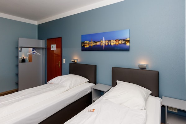 A hotel room with two single beds, a wardrobe, a mirror, a painting of a cityscape, and a nightstand with a lamp.