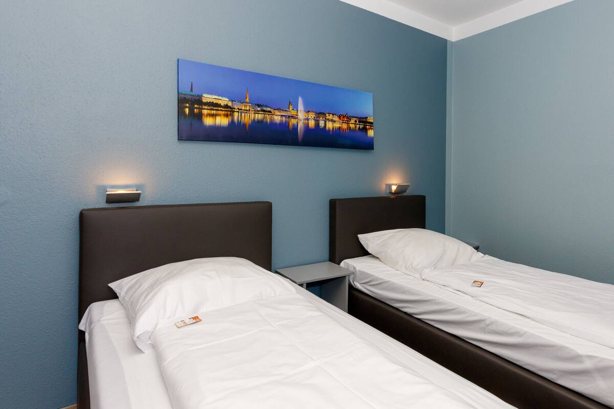 A hotel room with two single beds, a nightstand, a wall-mounted lamp, and a painting of a cityscape.