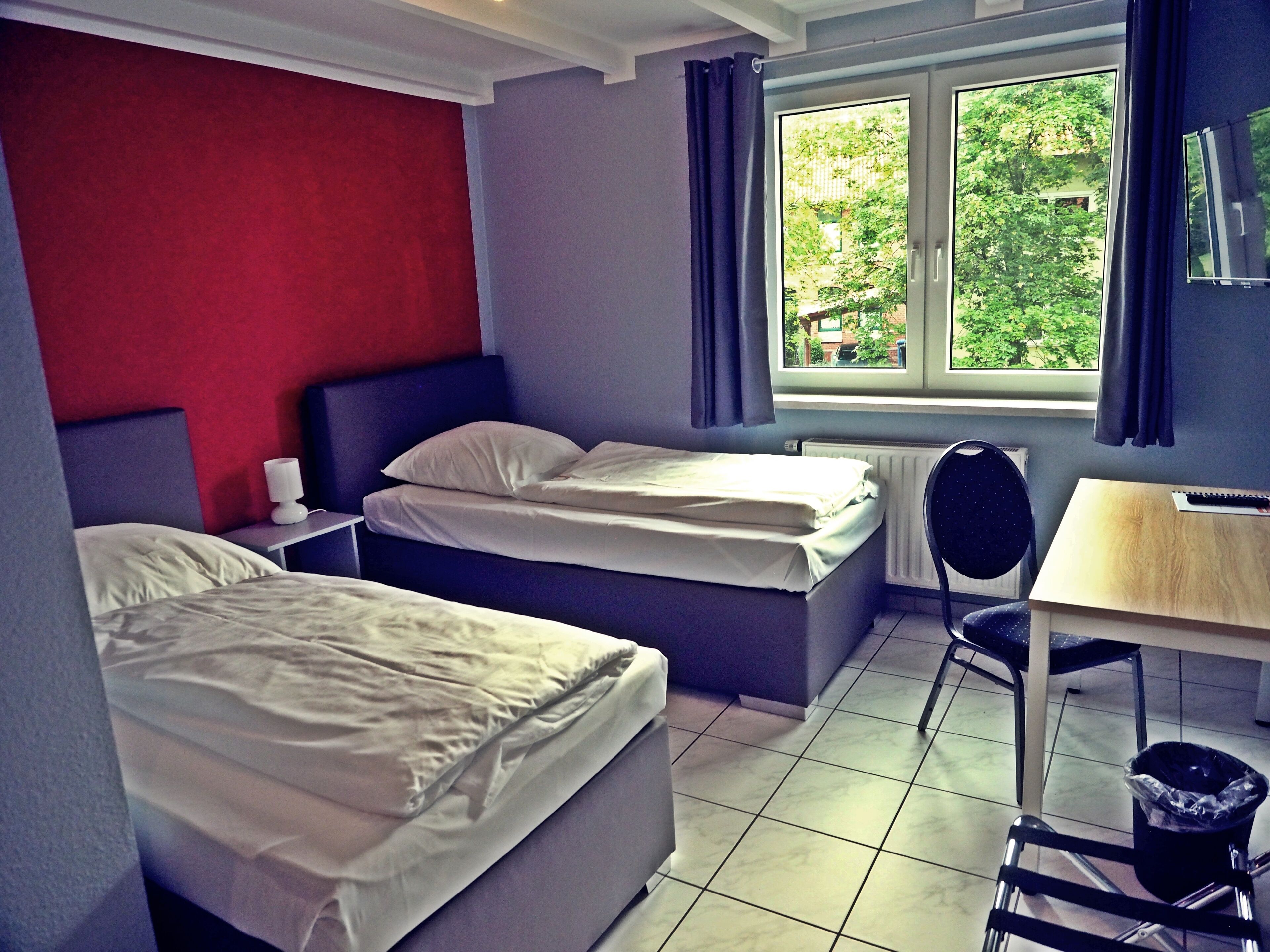 A hotel room with two beds, a desk, a chair, a radiator, and a window with curtains.