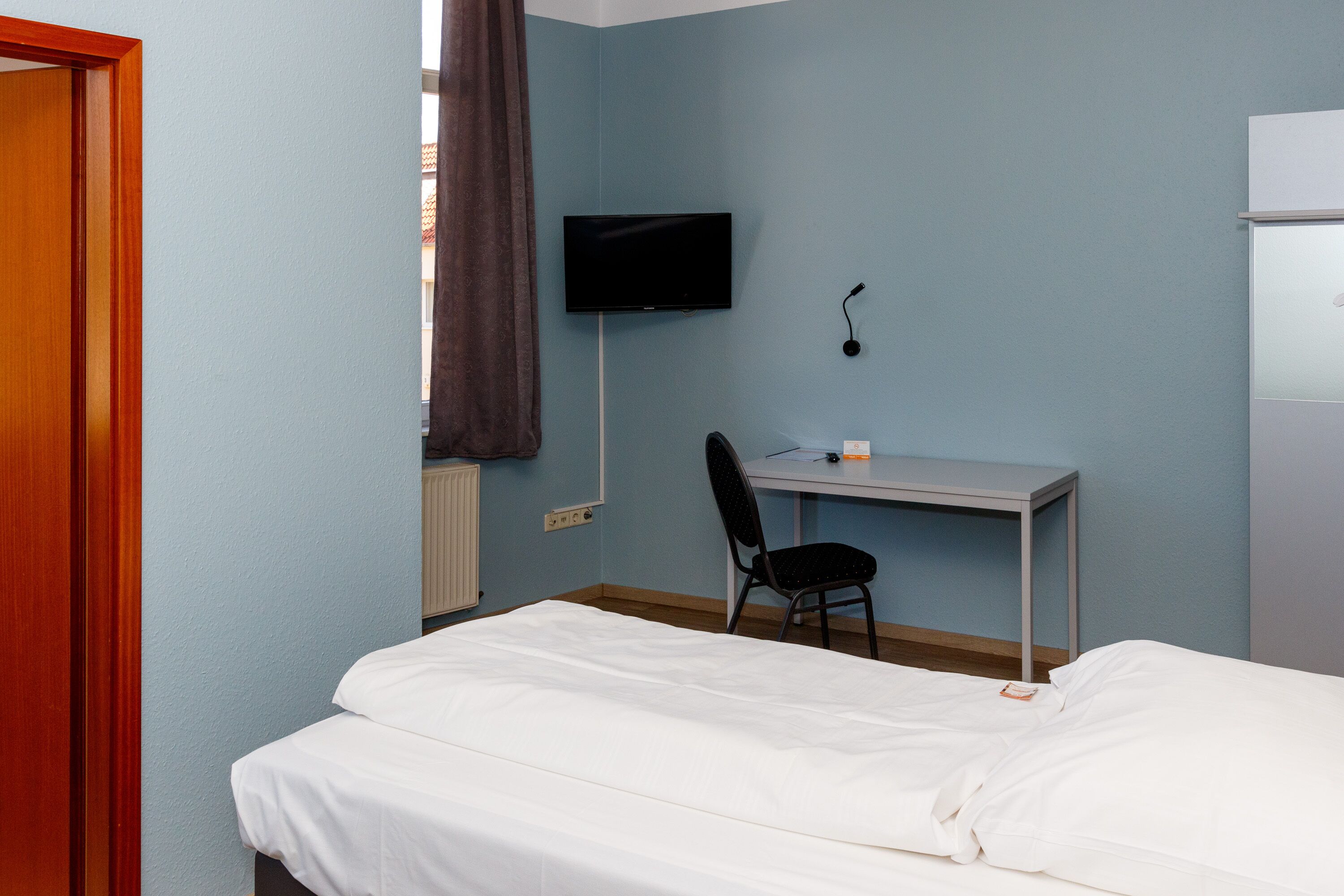 Een slaapkamer met een bed, een bureau, een stoel, een televisie en een raam met gordijnen.