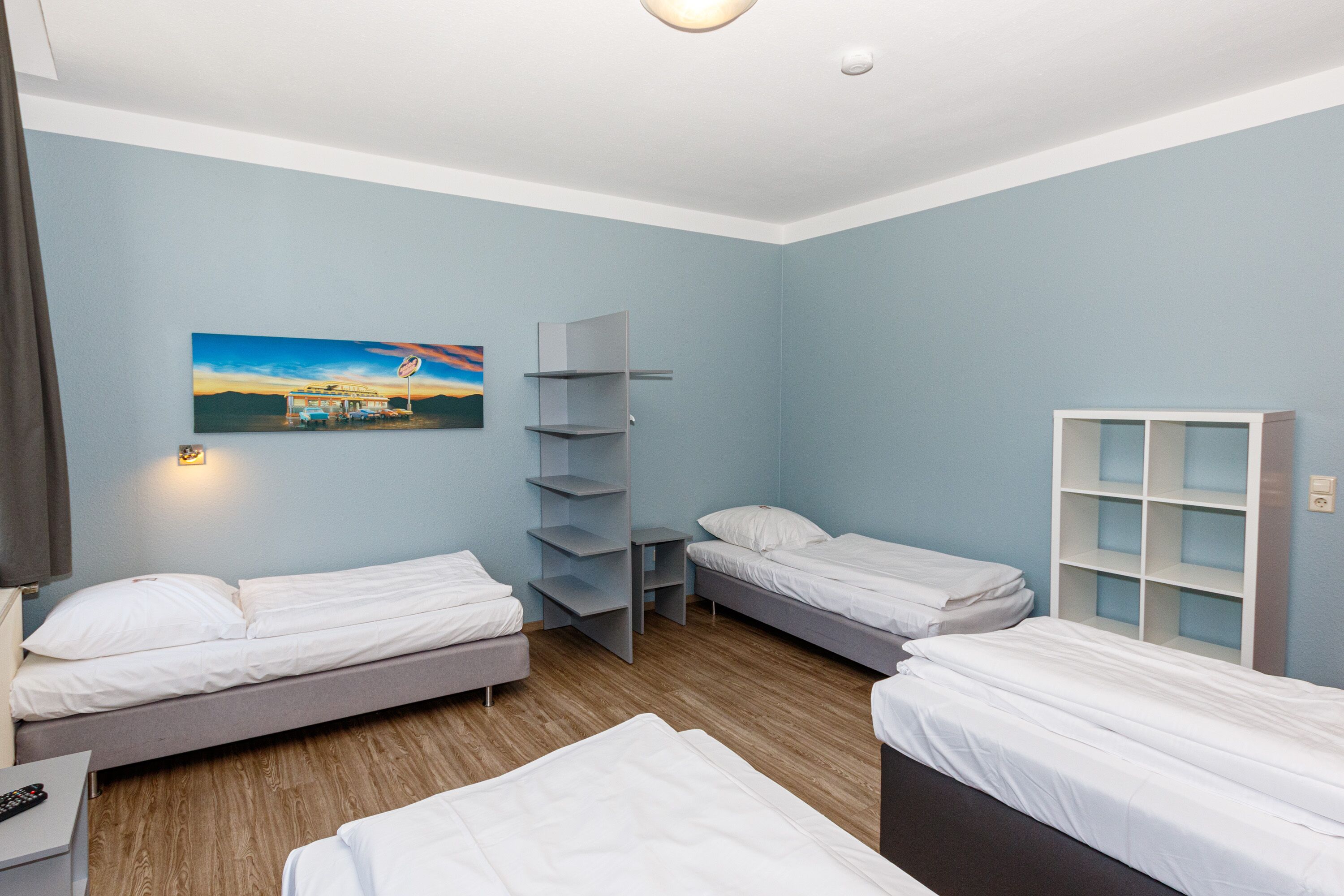 Habitación de hotel con dos camas individuales, un escritorio, una silla, una mesita y un estante de pared.
