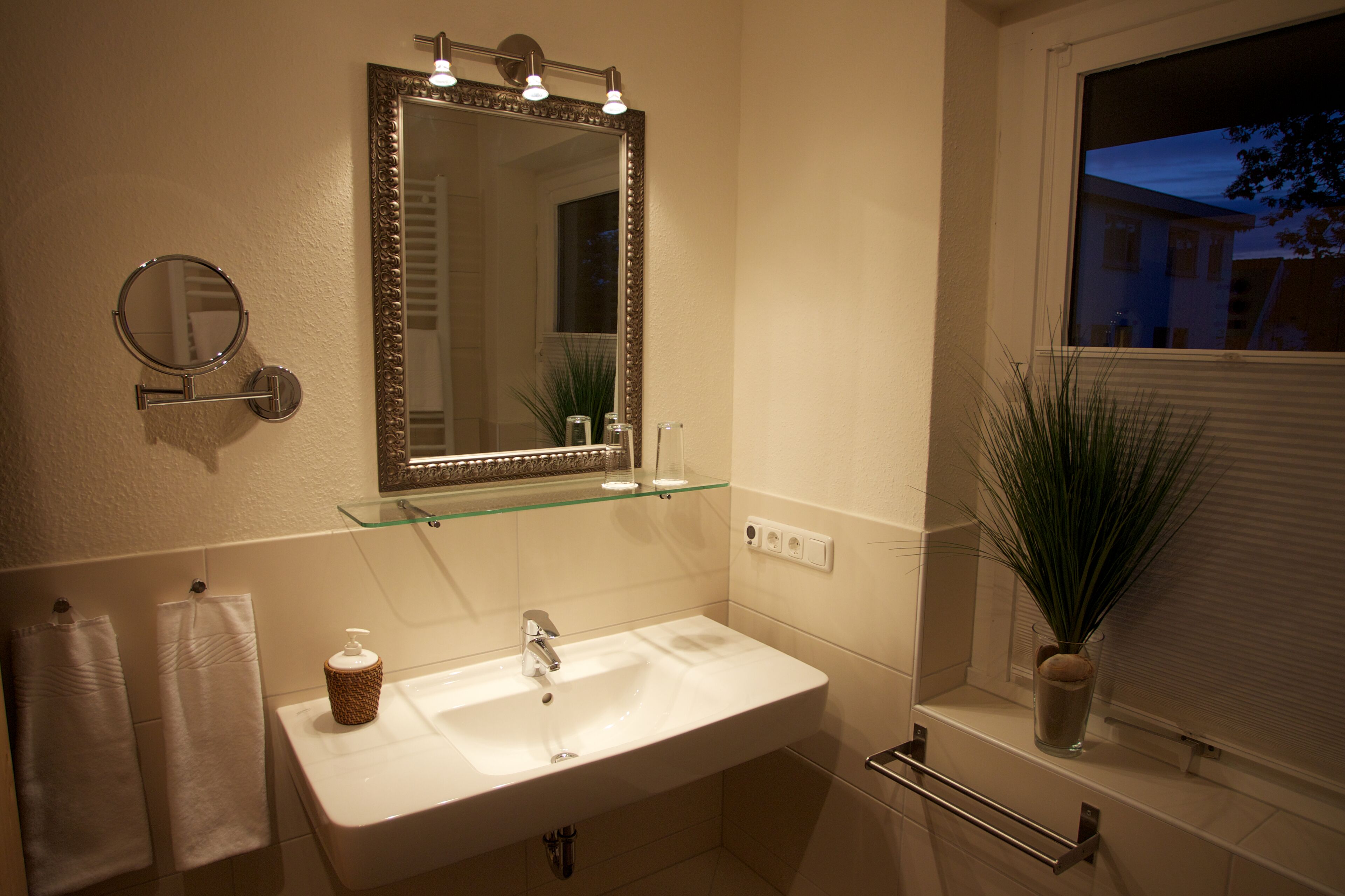 Une salle de bain avec un lavabo blanc, un miroir avec éclairage intégré, un porte-serviettes et une fenêtre avec des stores.