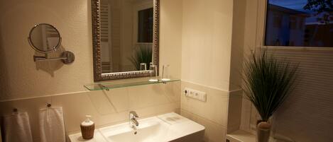 A bathroom with a white sink, a mirror with lights, a towel rack, and a window with blinds.