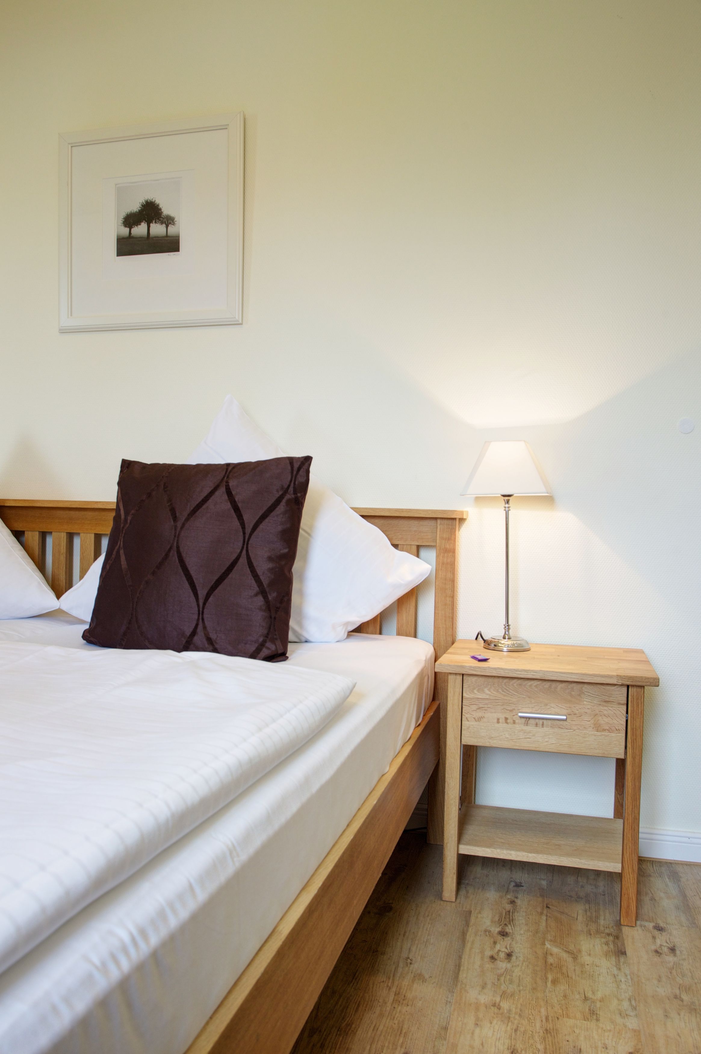 Een netjes opgemaakt bed met witte lakens en een bruin kussen, een nachtkastje met een lamp en een lijst met een foto aan de muur.