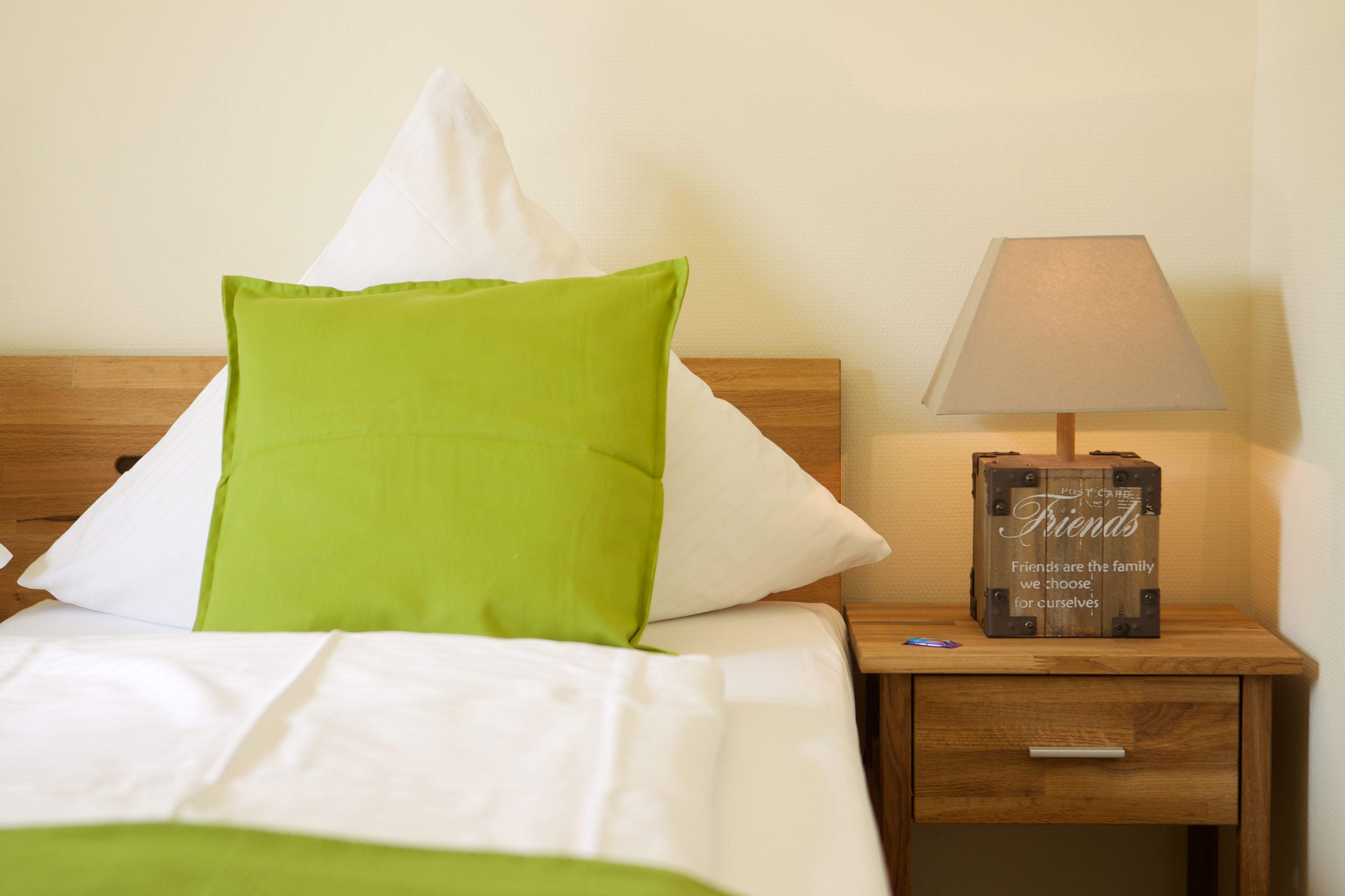 Un lit bien fait, avec des oreillers blancs et verts, une lampe de chevet et une table de chevet en bois sur laquelle se trouve une boîte décorative.