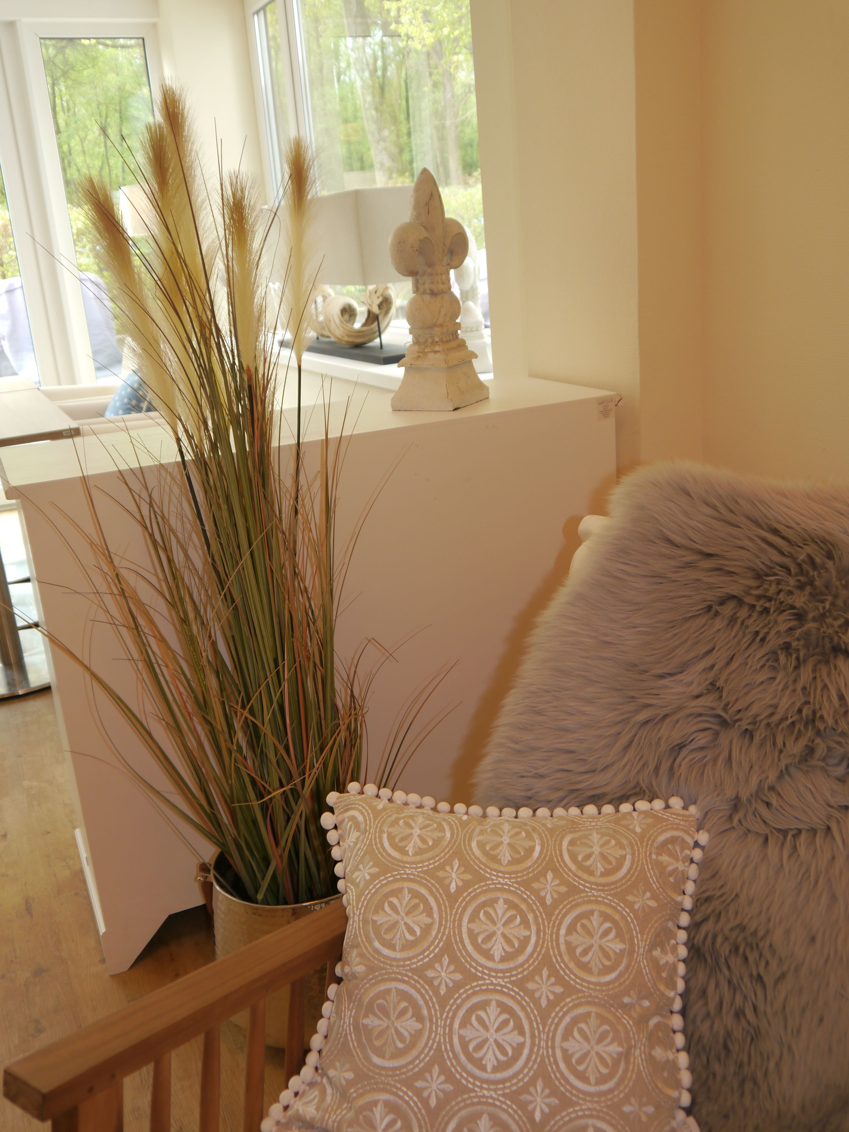Una habitación con un escritorio blanco, una planta en maceta con hierbas altas, un jarrón blanco decorativo y un almohadón gris suave.