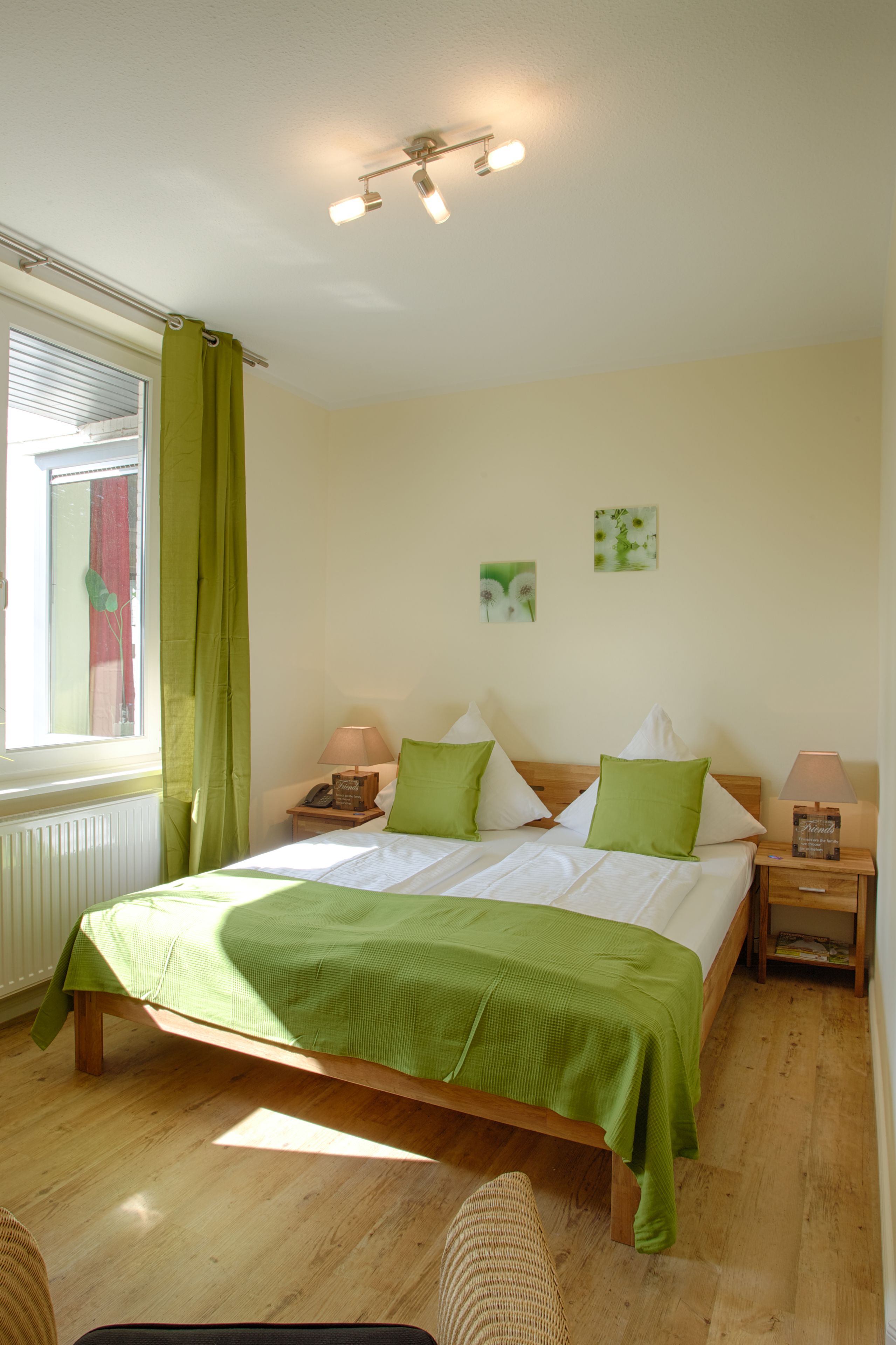 Une chambre à coucher avec un lit en bois, une literie verte, deux tables de chevet avec des lampes, une fenêtre avec des rideaux et des œuvres d’art accrochées au mur.
