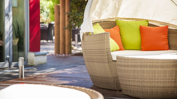 A wicker sofa with a canopy, orange and green cushions, and a metal bollard in a courtyard.