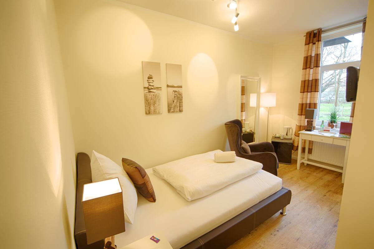 Une chambre d’hôtel avec un lit, un bureau et une chaise. Il y a deux tableaux accrochés au mur et une fenêtre avec des rideaux.