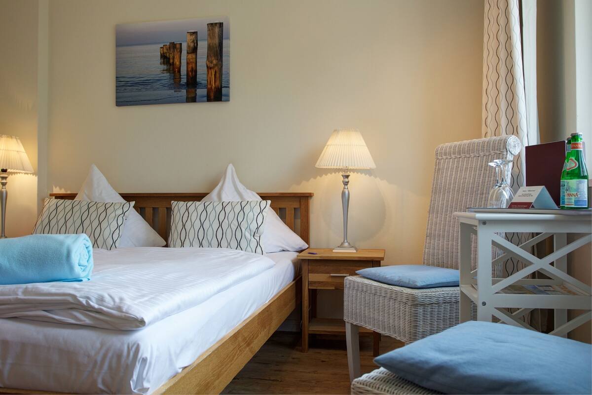 Un lit bien fait, avec du linge de lit blanc et une serviette bleue, une chaise en osier avec un coussin, une petite table avec une lampe et un tableau encadré au mur.