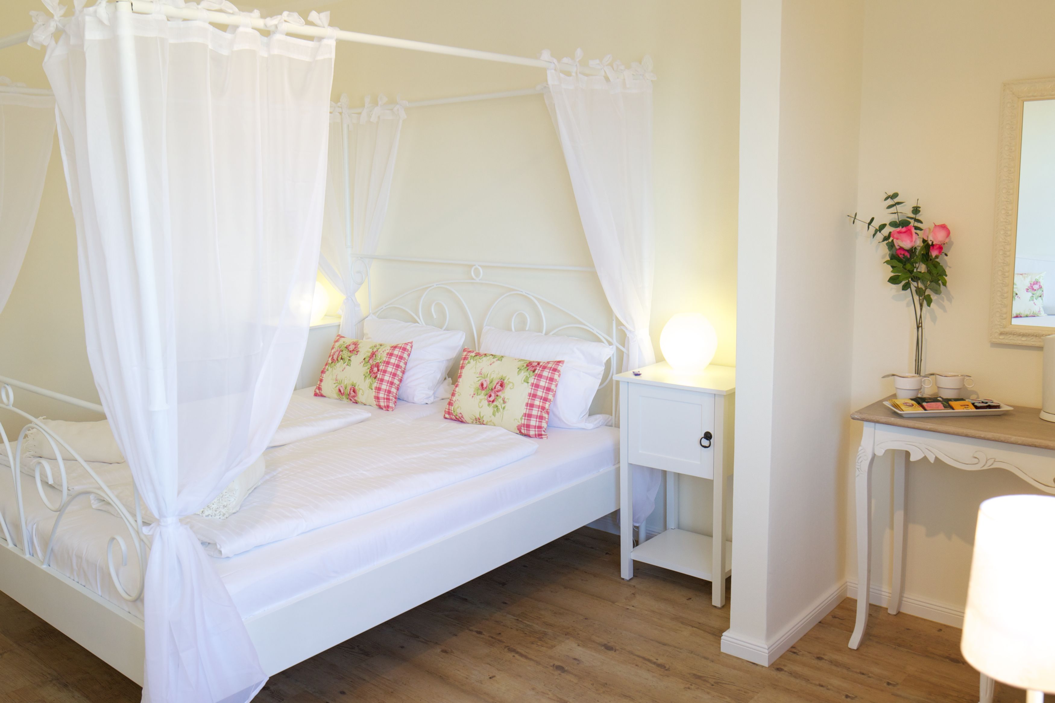A four-poster bed with white bedding and floral pillows, a bedside table with a lamp, and a mirror on an adjacent wall.