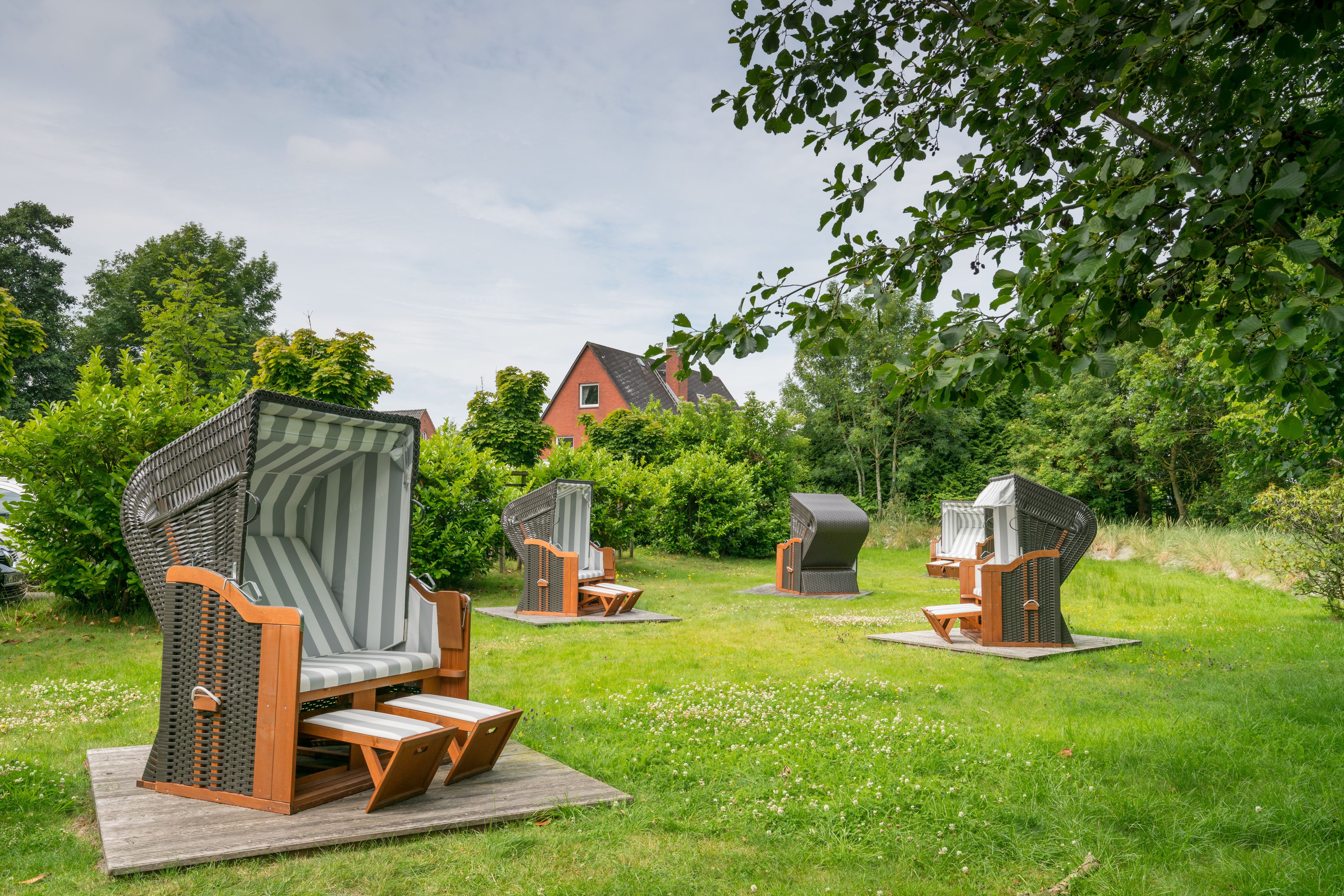 Outdoor seating with high backs and wooden legs, arranged on a grassy area with trees and a building in the background.