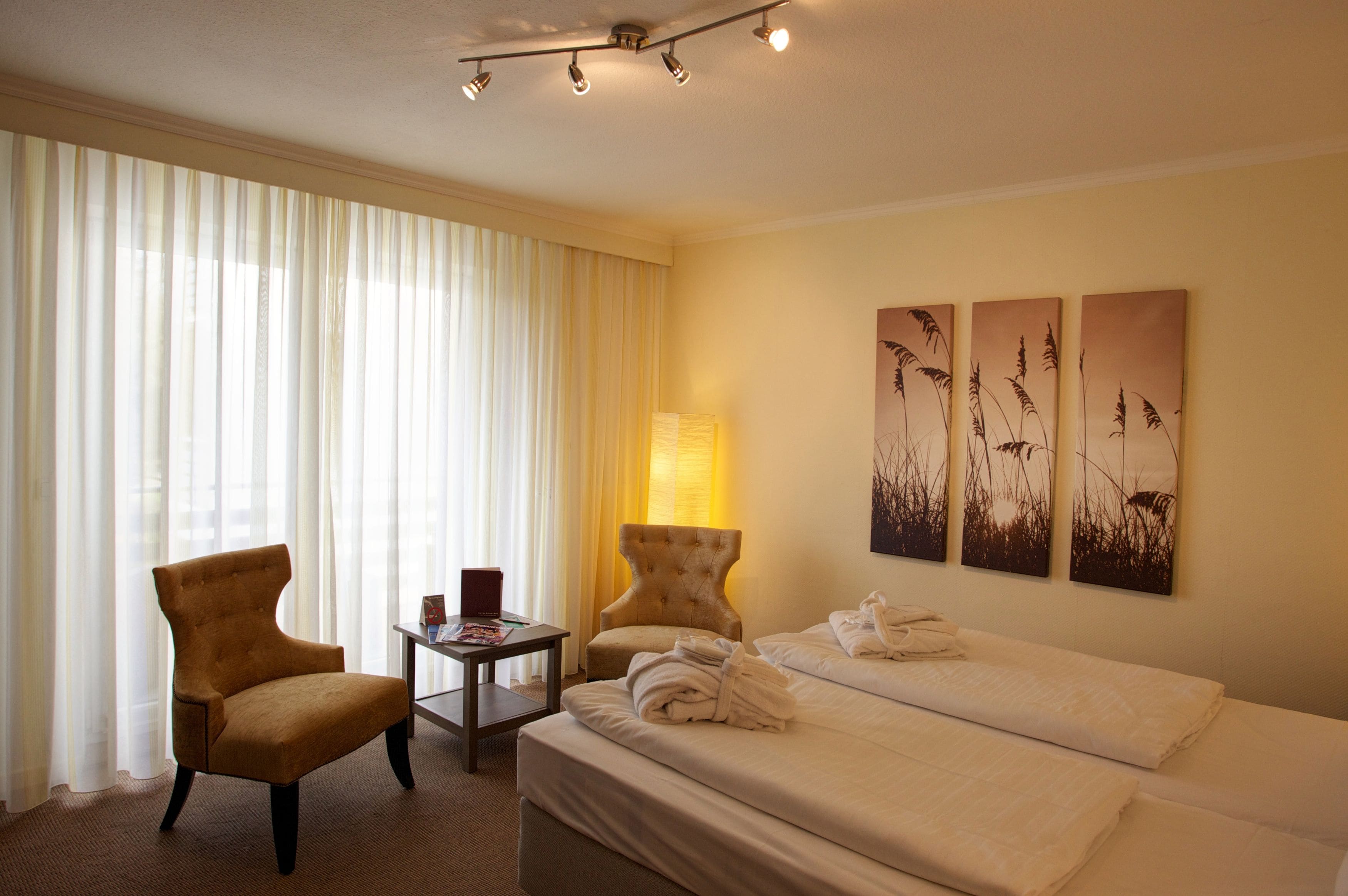 A hotel room with a bed, two armchairs, a side table, and a lamp. There are three wall-mounted paintings of grass.