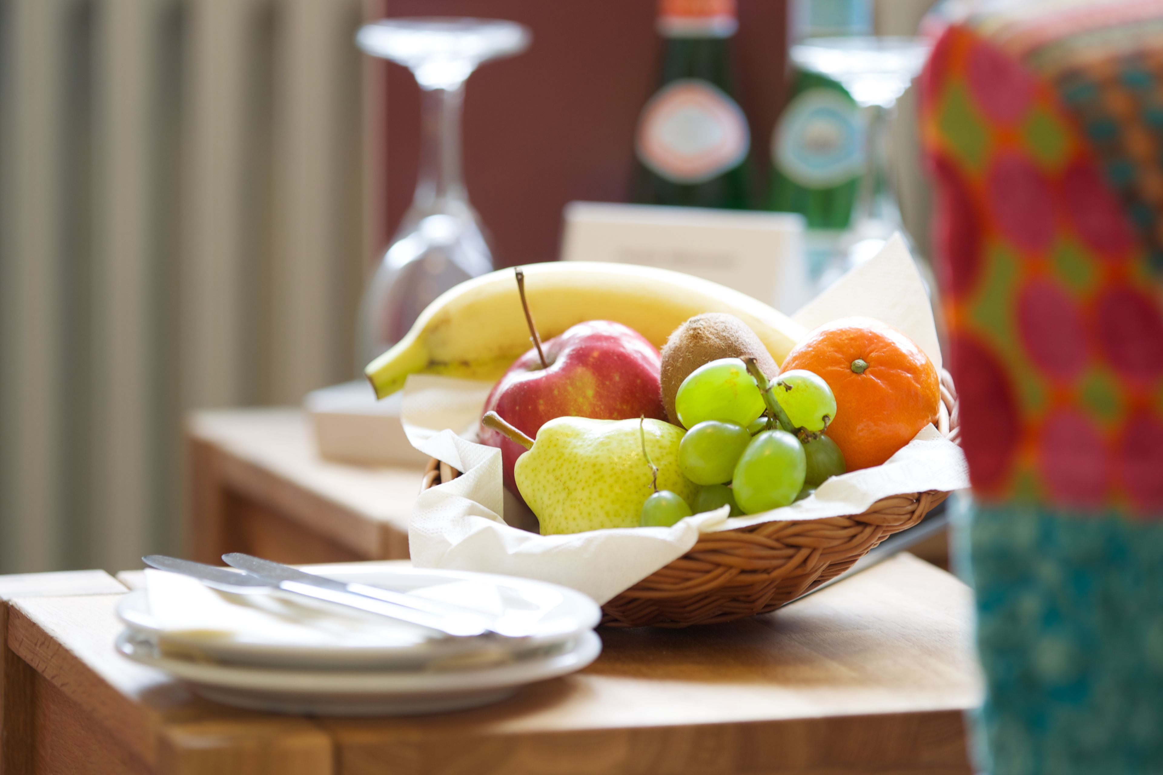 A fruit basket on a table with a wine glass and a bottle in the background.