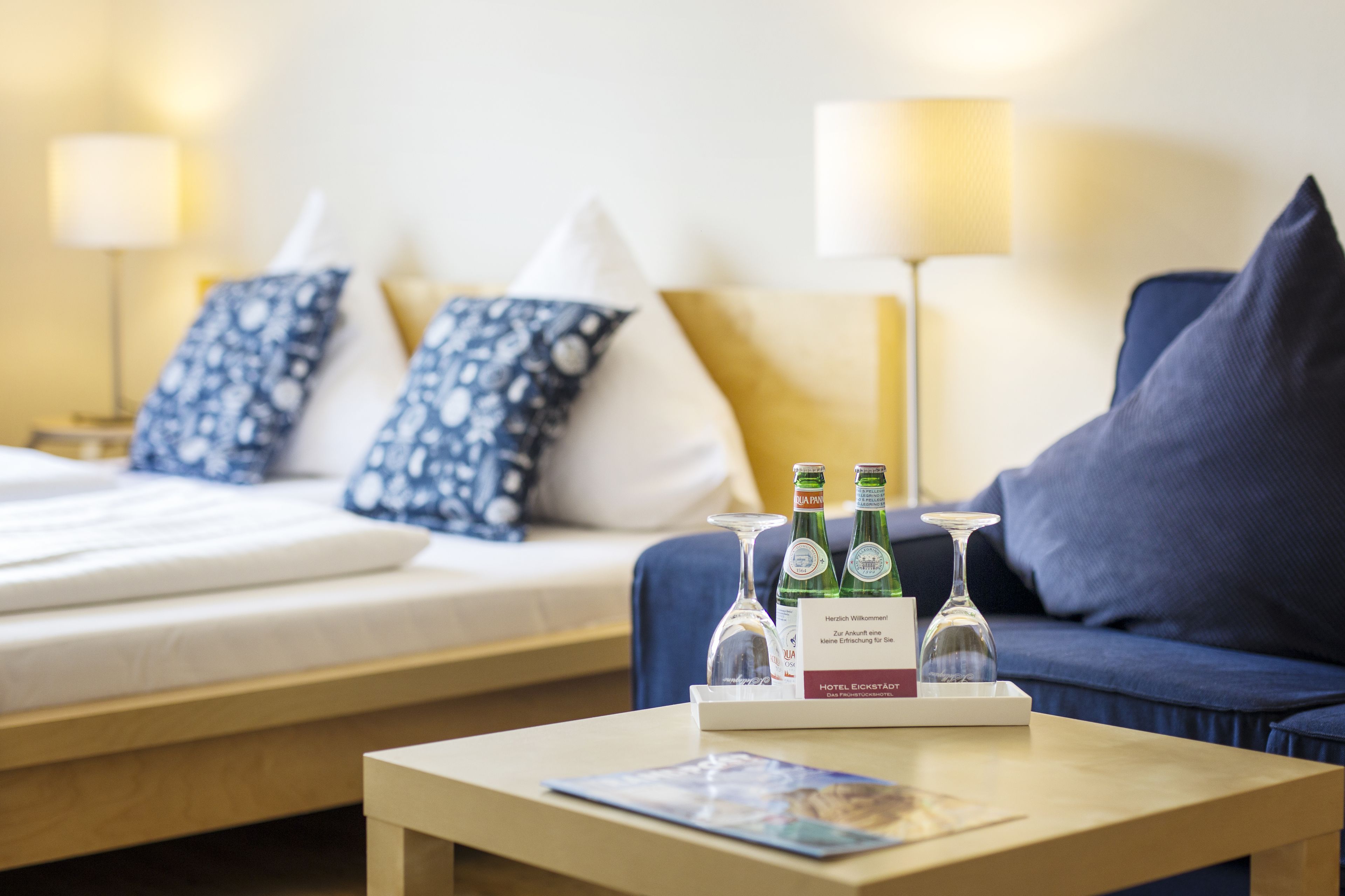 Quarto de hotel com uma cama, um sofá azul, uma mesa com uma bandeja de garrafas e copos, e um abajur.