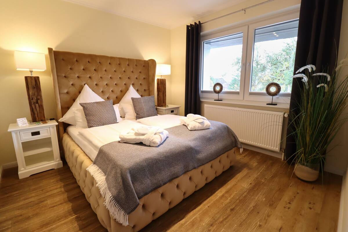 Une chambre à coucher avec une tête de lit capitonnée, un grand lit recouvert d’une couverture à rayures grises et blanches, deux lampes de chevet et une fenêtre avec des rideaux.