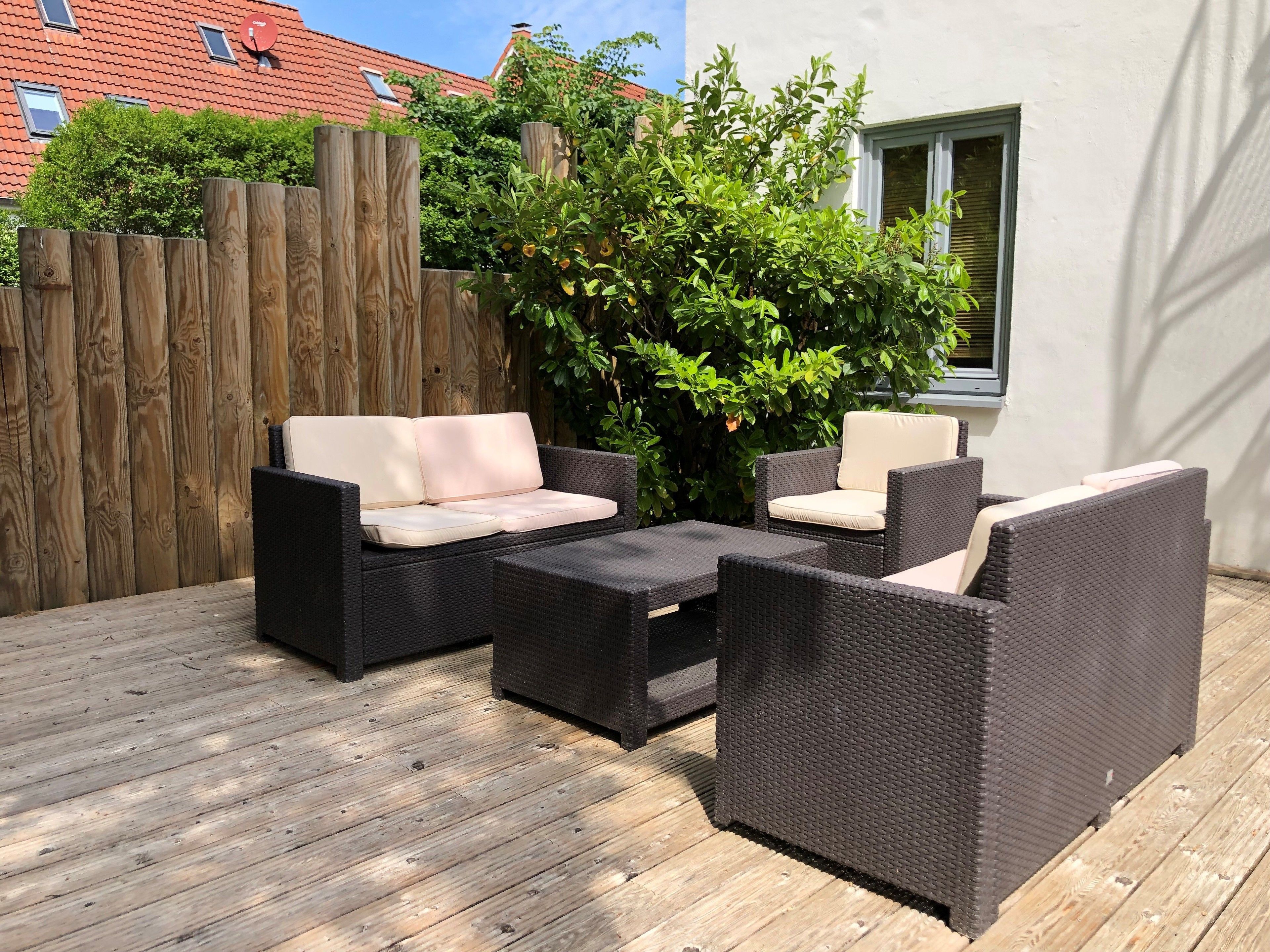 Un patio al aire libre con mobiliario de mimbre, una terraza de madera y un fondo de edificio y follaje.