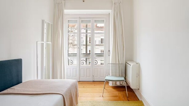 Une chambre à coucher avec un lit, une chaise, un radiateur et une vue sur l’extérieur, visible à travers une porte.