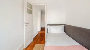 Une chambre avec un lit simple, une porte donnant sur un couloir et un plancher en bois.