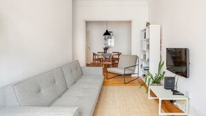 Un salon moderne avec un canapé gris, une table à manger avec des chaises, un téléviseur posé sur un meuble et une bibliothèque.