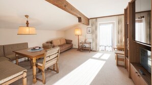 A cozy living room with a wooden ceiling, a dining table with chairs, a sofa, a TV, and a lamp. - Hotel Bernhard am See (Walchsee)