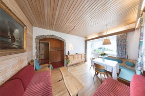 A cozy room with a wooden ceiling, a stone fireplace, a dining area with a table and chairs, and a large window with curtains. - Tirolerhaus Walchsee (Walchsee)