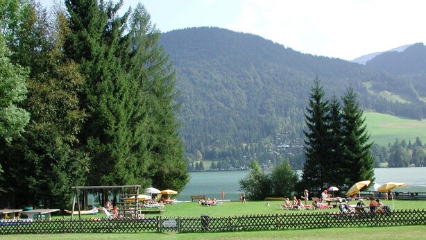 Vista de un lago con un área verde, mesas de picnic y sombrillas.