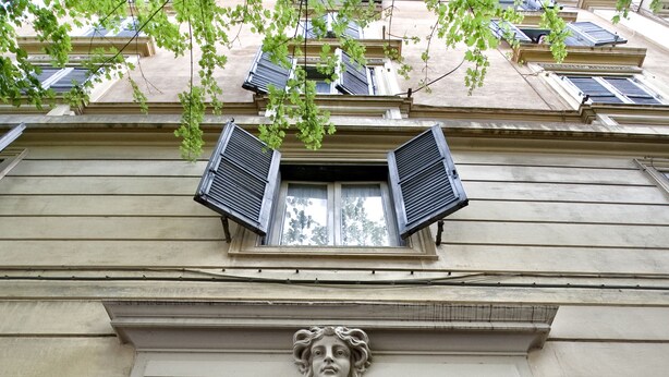 Une entrée d’immeuble ornée d’un bas-relief en pierre au-dessus de la porte, d’une petite fenêtre avec volets et d’une sonnette.