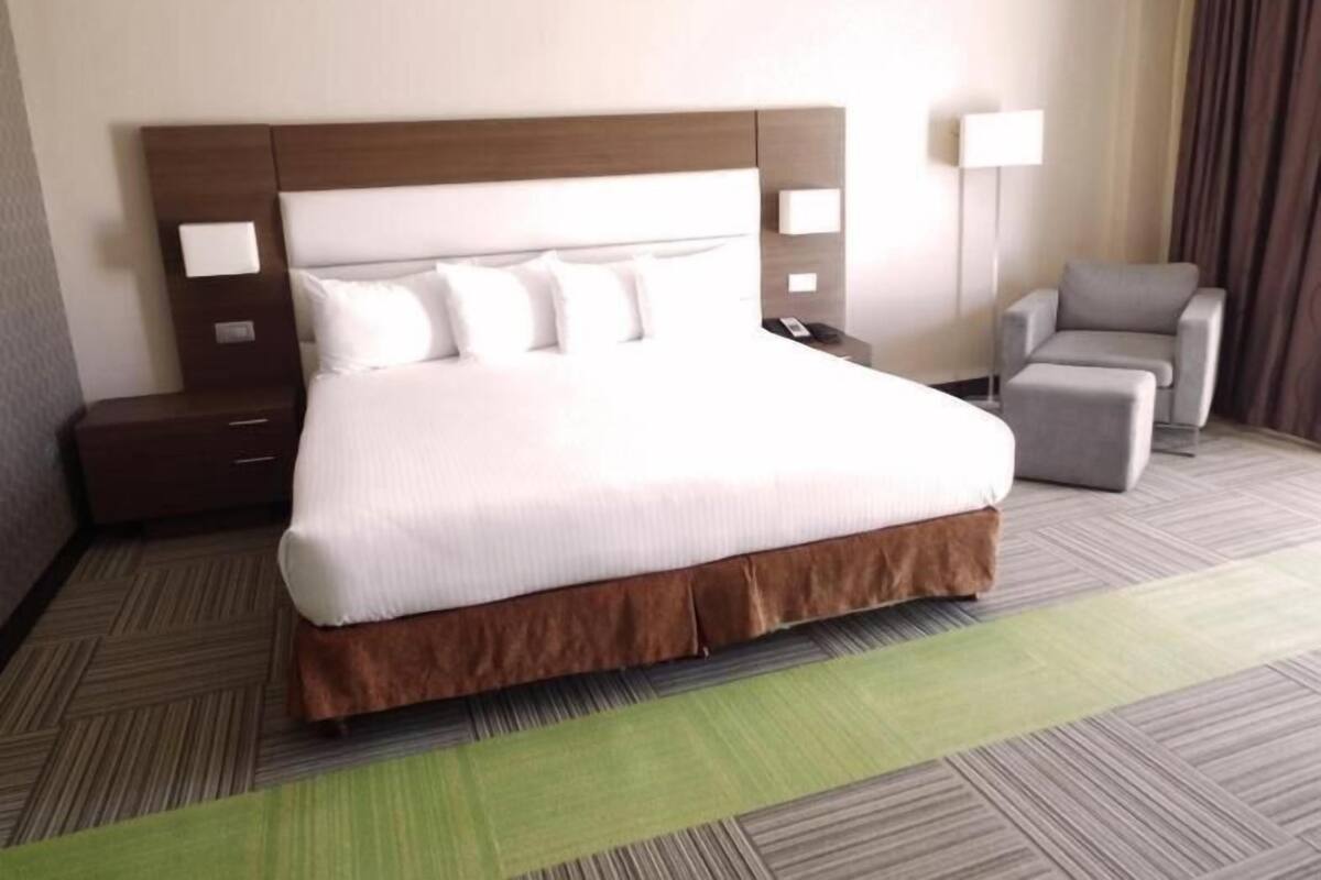 Una habitación de hotel con una cama grande, una mesita de noche, una silla y una lámpara.