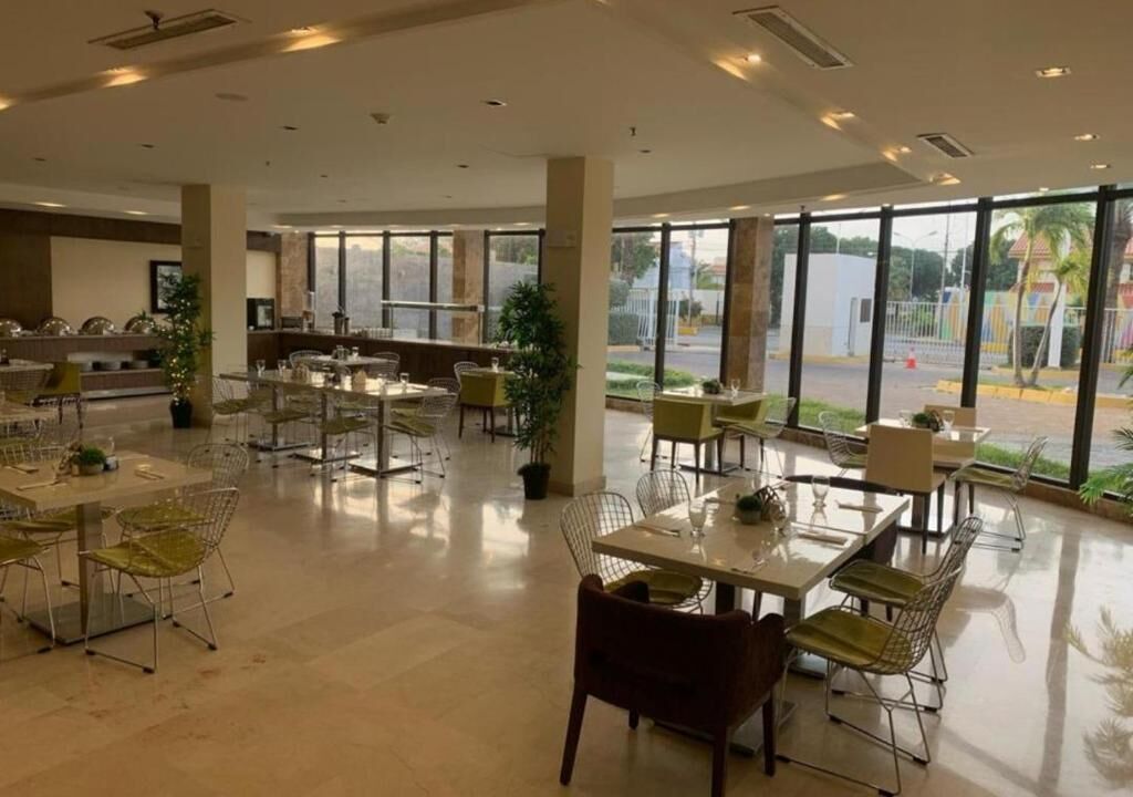 A spacious indoor area with multiple tables and chairs, large windows offering a view of the outside, and a reception desk in the background.