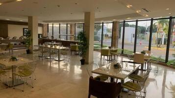 A spacious indoor area with multiple tables and chairs, large windows offering a view of the outside, and a reception desk in the background.
