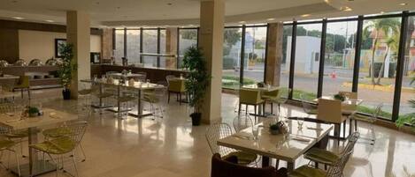 A spacious indoor area with multiple tables and chairs, large windows offering a view of the outside, and a reception desk in the background.