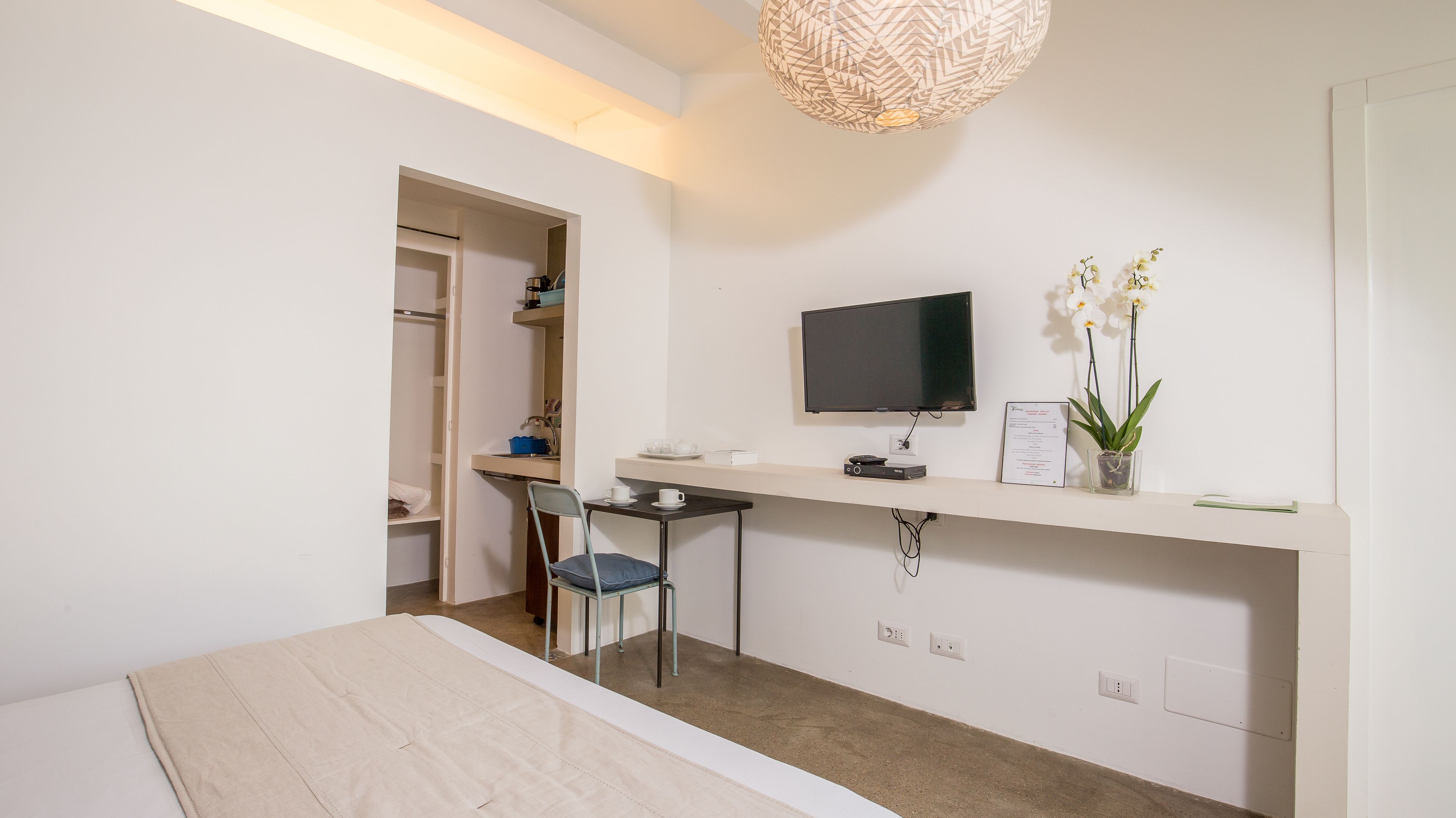 Quarto moderno e minimalista com uma escrivaninha de madeira, uma cadeira, uma televisão de tela plana, uma pequena orquídea em vaso e uma porta branca que dá para um armário.