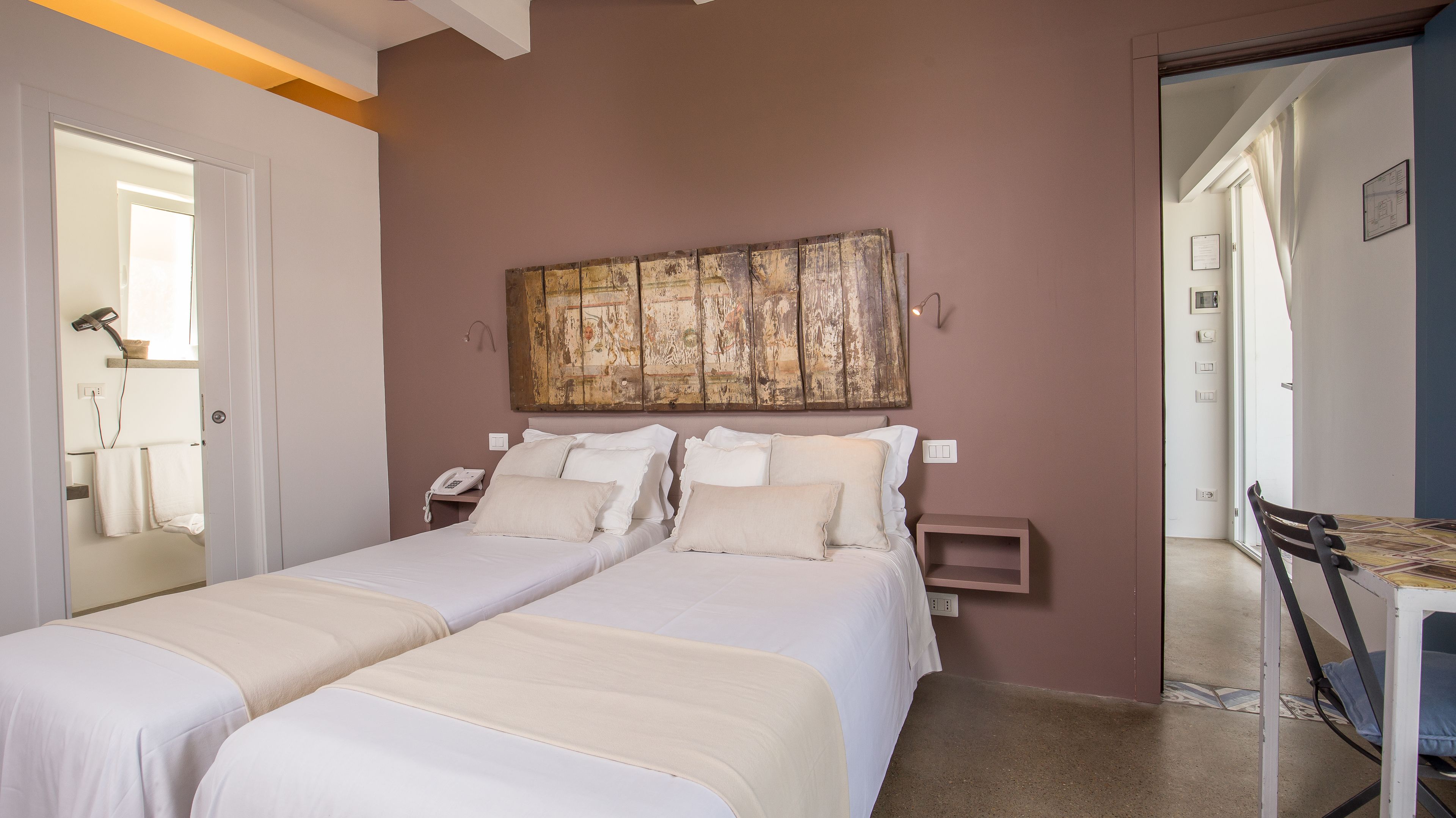 A bedroom with two beds, a wooden wall art piece, a bedside table with a lamp, and a chair.