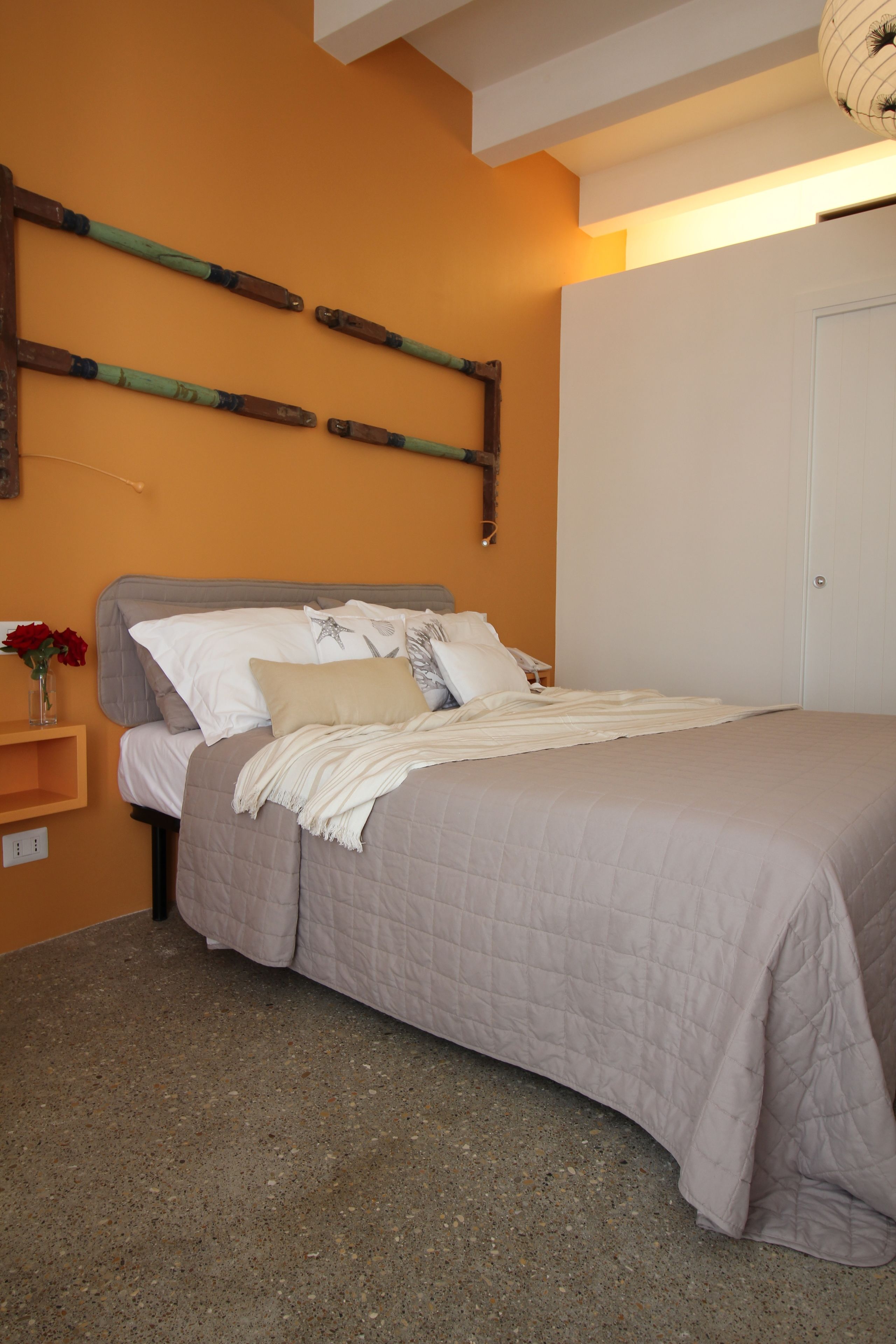 Un dormitorio con cama, almohadas, una mesita de noche con un florero y una estantería de pared.