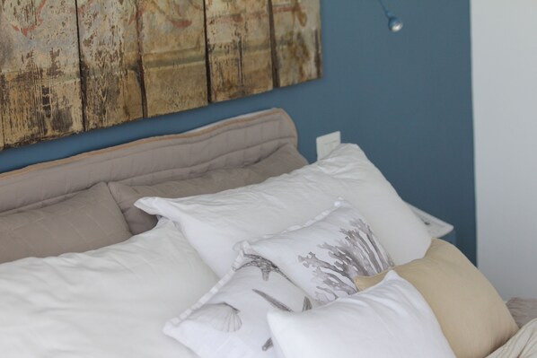 A bed with white and beige bedding, a blue wall, and a wooden artwork above it.