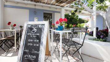 Un patio de hotel con mesas al aire libre, un cartel de pizarra con precios de habitaciones y plantas en macetas.