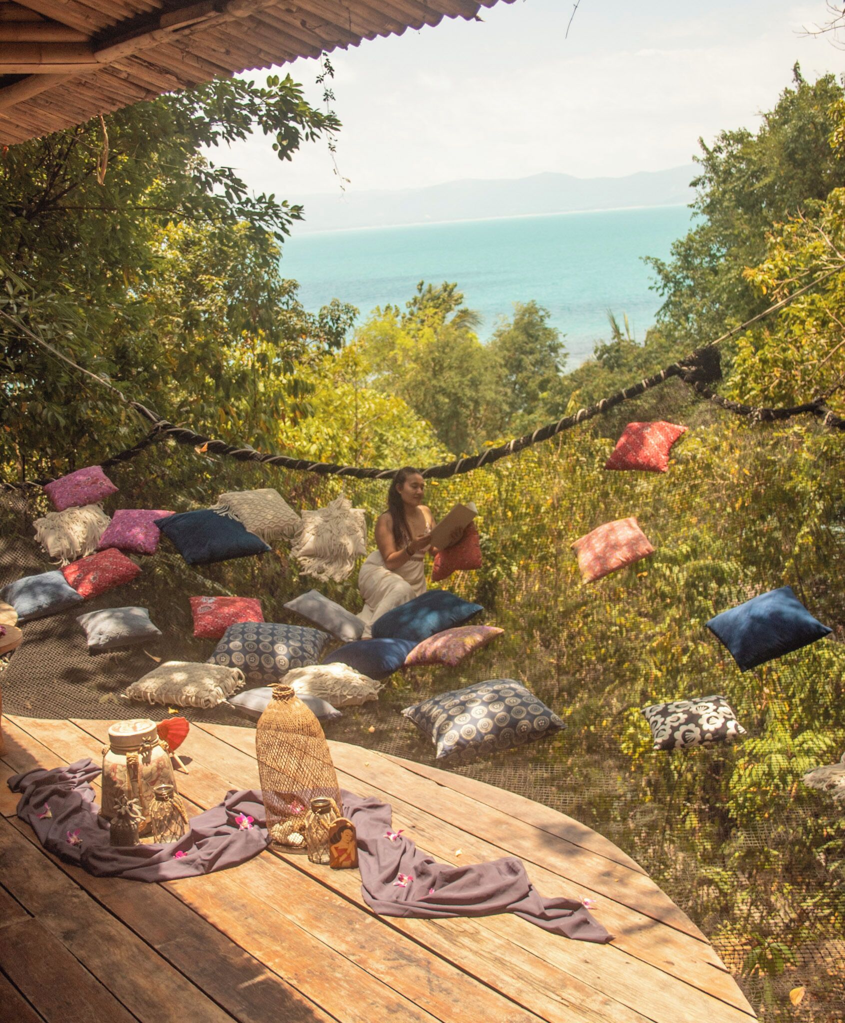 Terraza de madera con vistas al mar, rodeada de vegetación y una hamaca con almohadones.