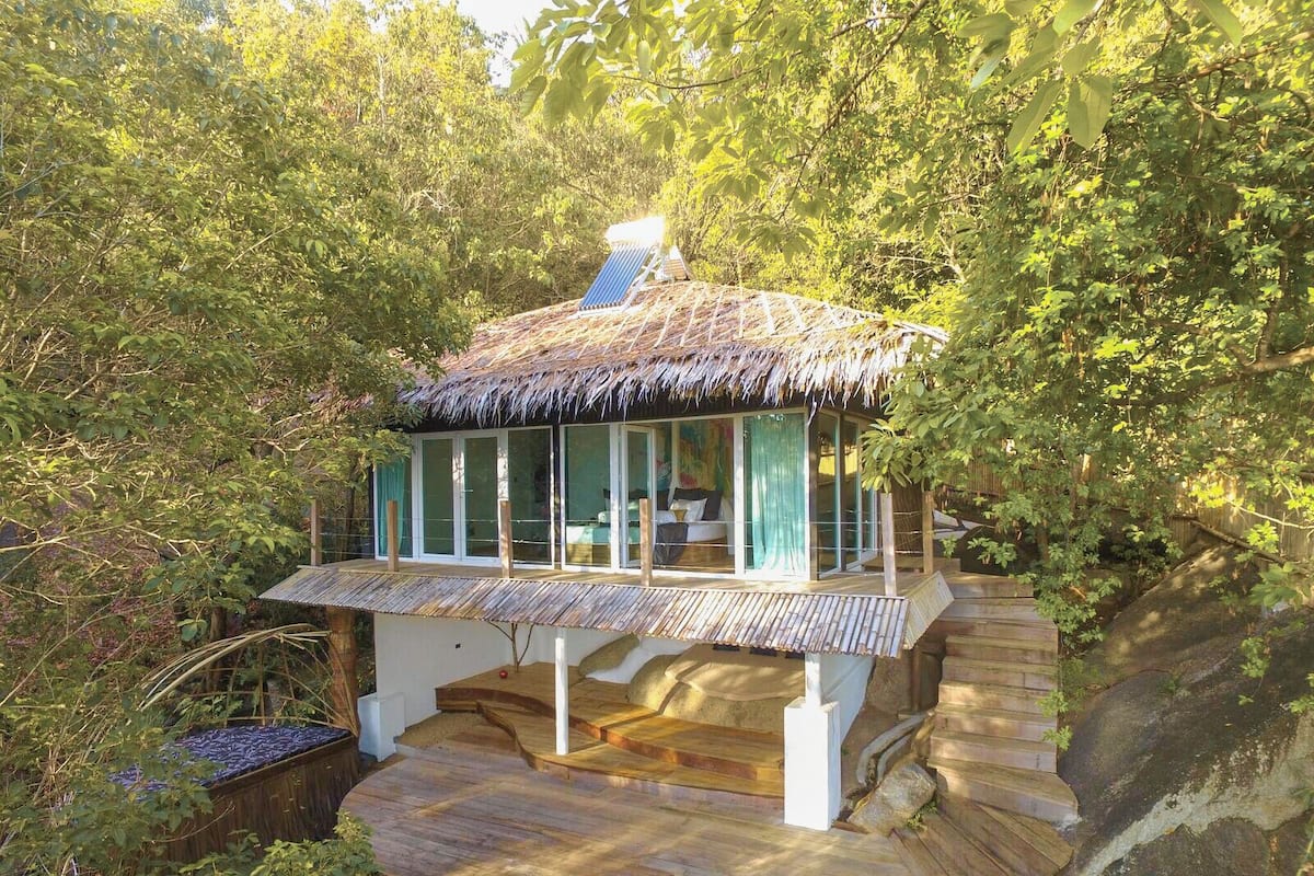 A thatched-roof structure with a glass enclosure, surrounded by dense foliage.
