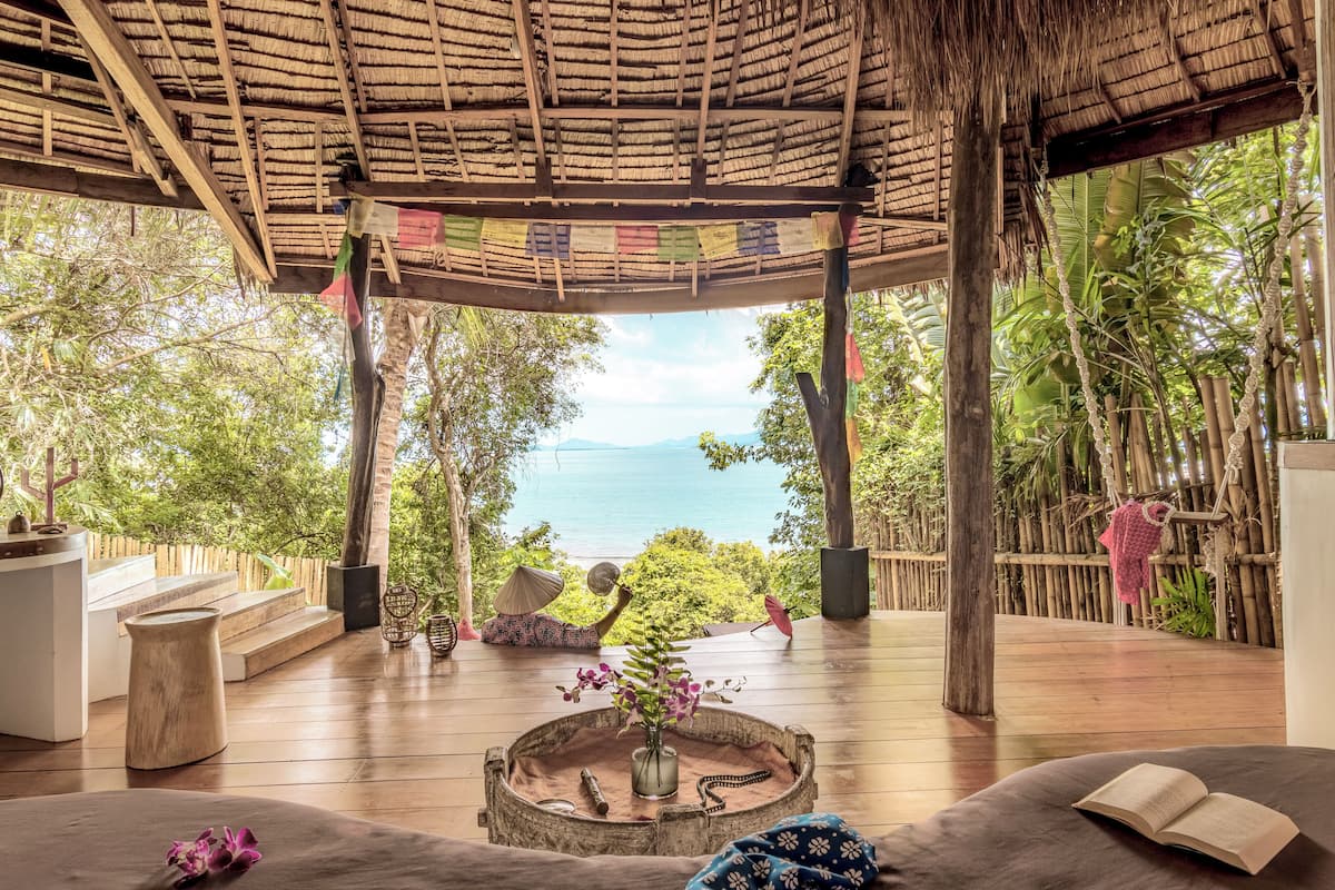 A wooden deck with a thatched roof, a view of the sea, and a seating area with a book and flowers.