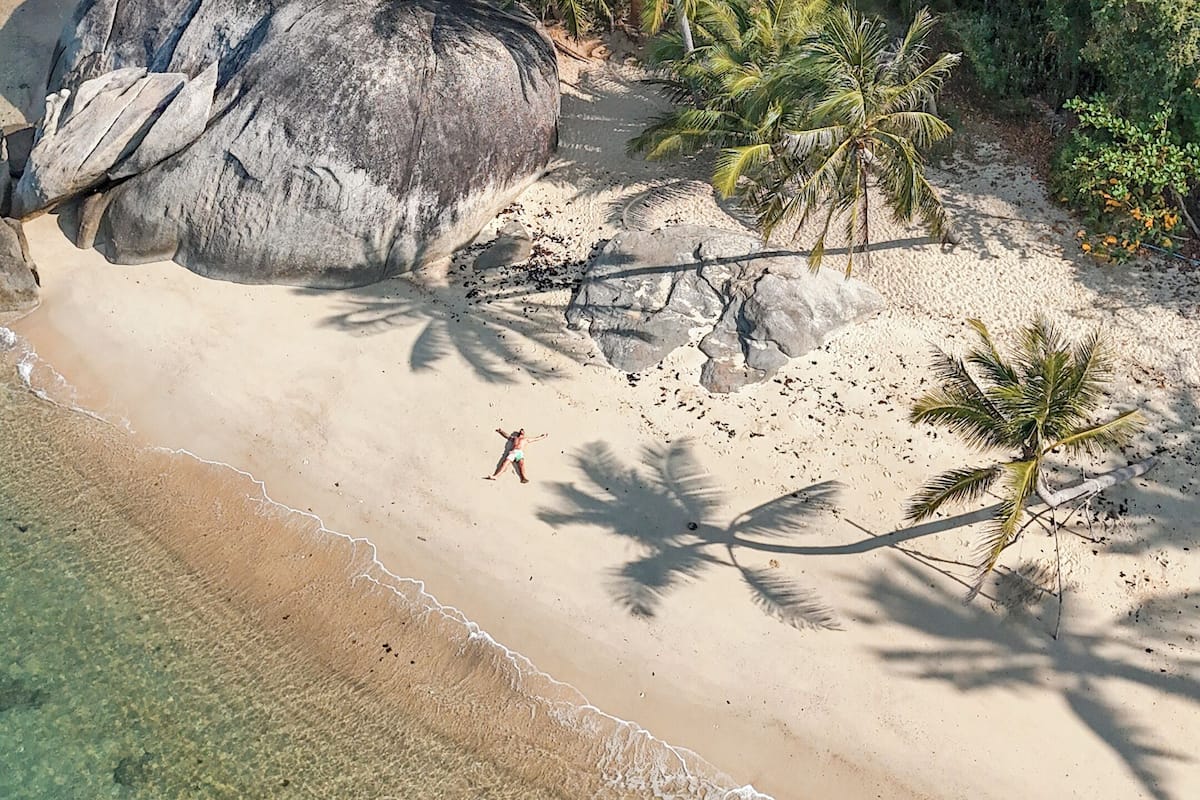 A beach with clear water, a large rock, and a house surrounded by dense greenery.