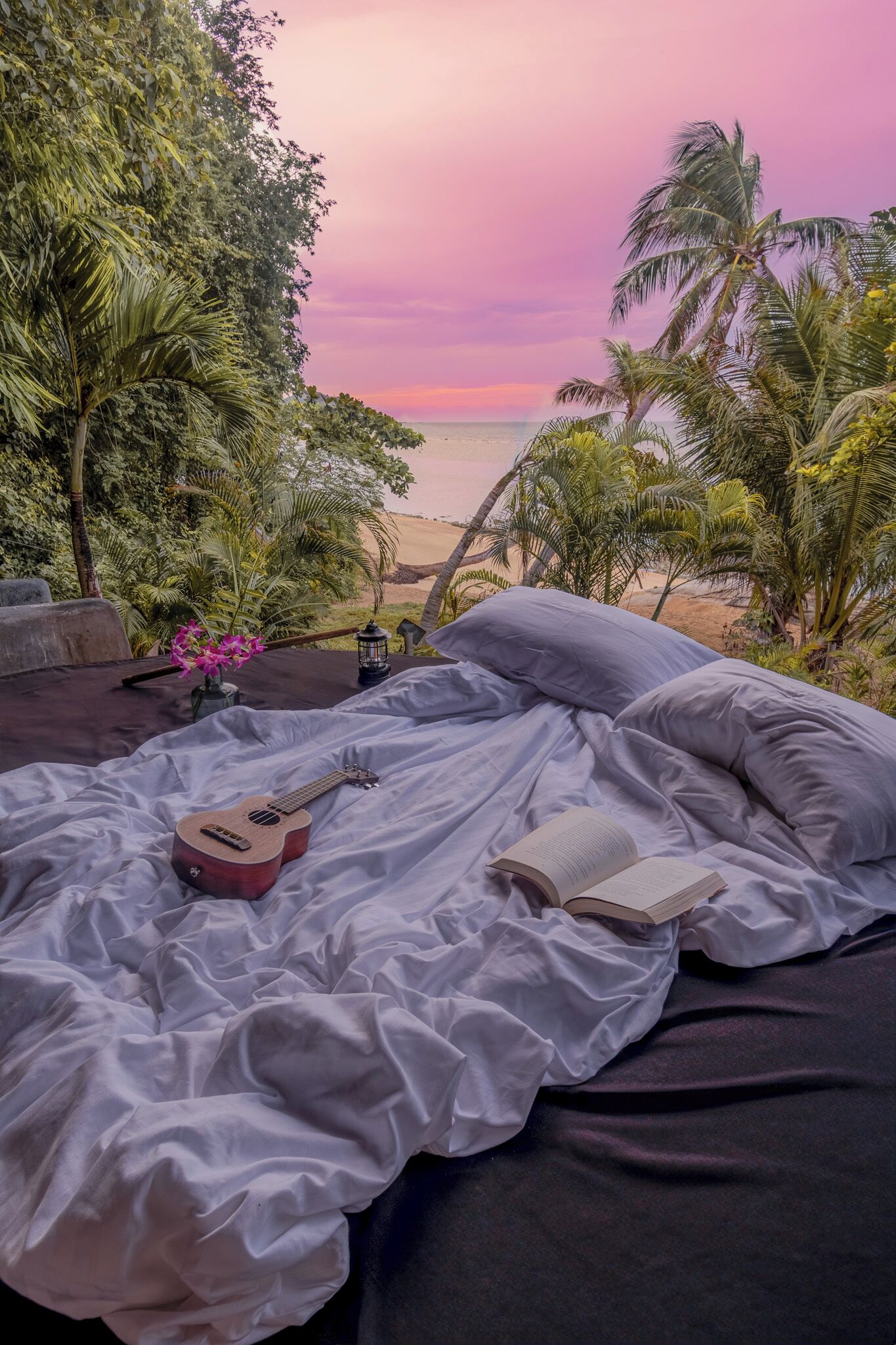 Una cama con una guitarra y un libro, frente a una vista a la playa con palmeras.