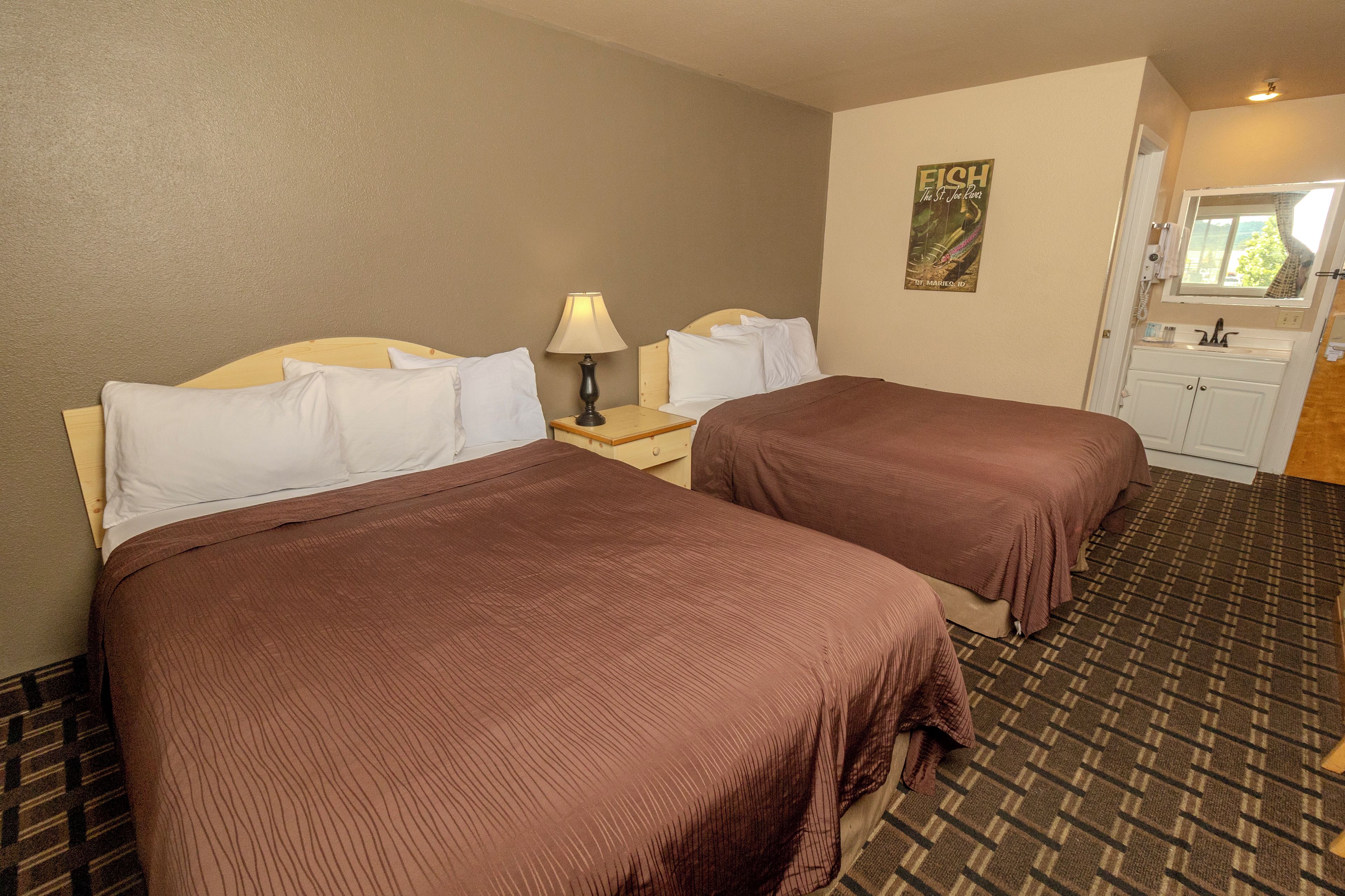 A hotel room with two beds, a nightstand with a lamp, a painting on the wall, and a bathroom visible through an open door.