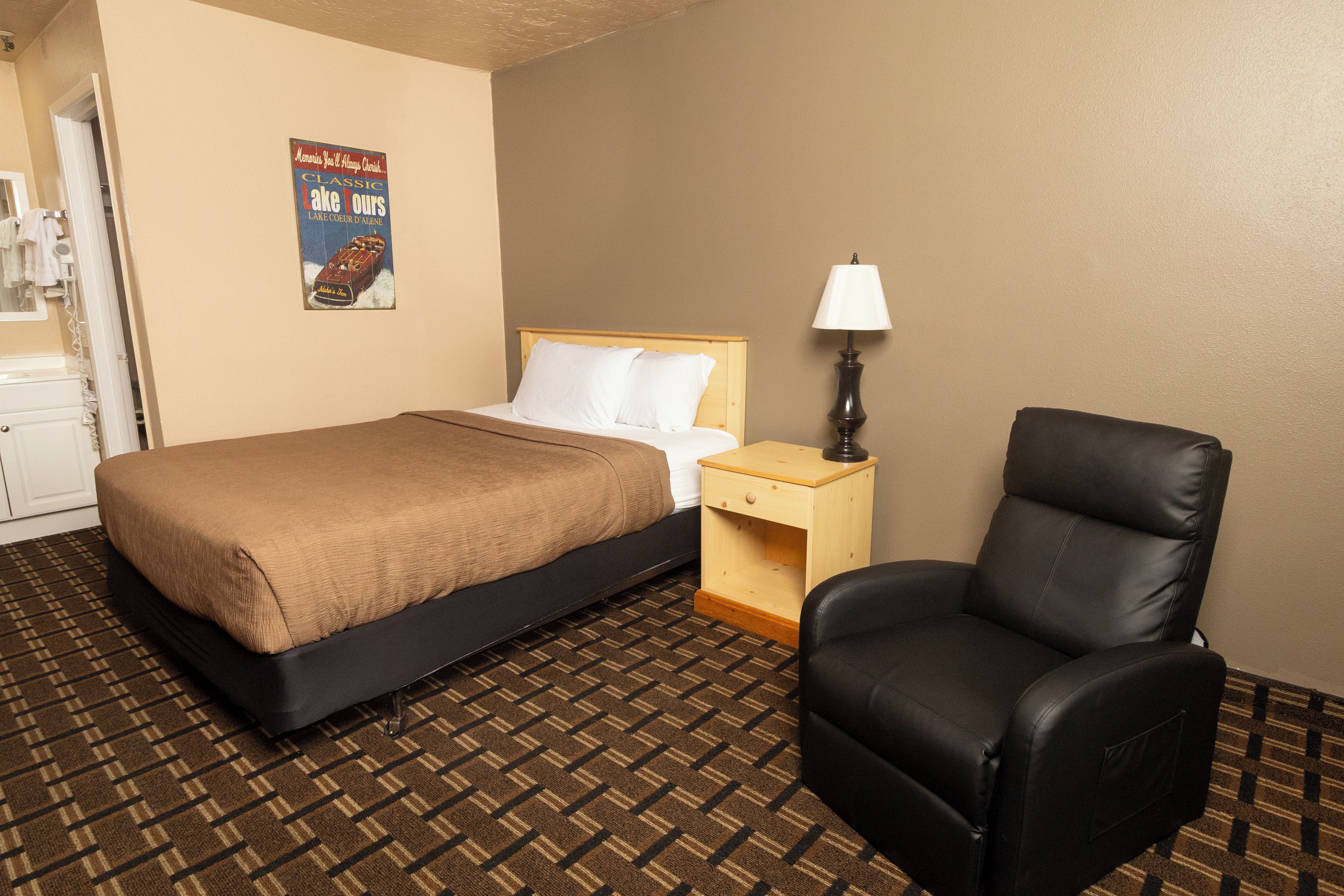A hotel room with a bed, a chair, a nightstand, a lamp, and a poster on the wall.