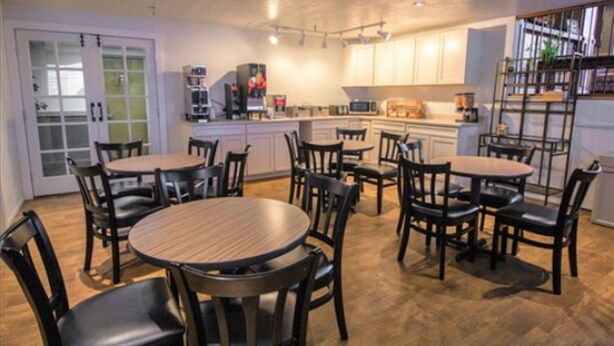 A modern kitchen with white cabinets, a central island, and a dining area with round tables and chairs. - FairBridge Inn - Coeur d'Alene (Coeur d'Alene)