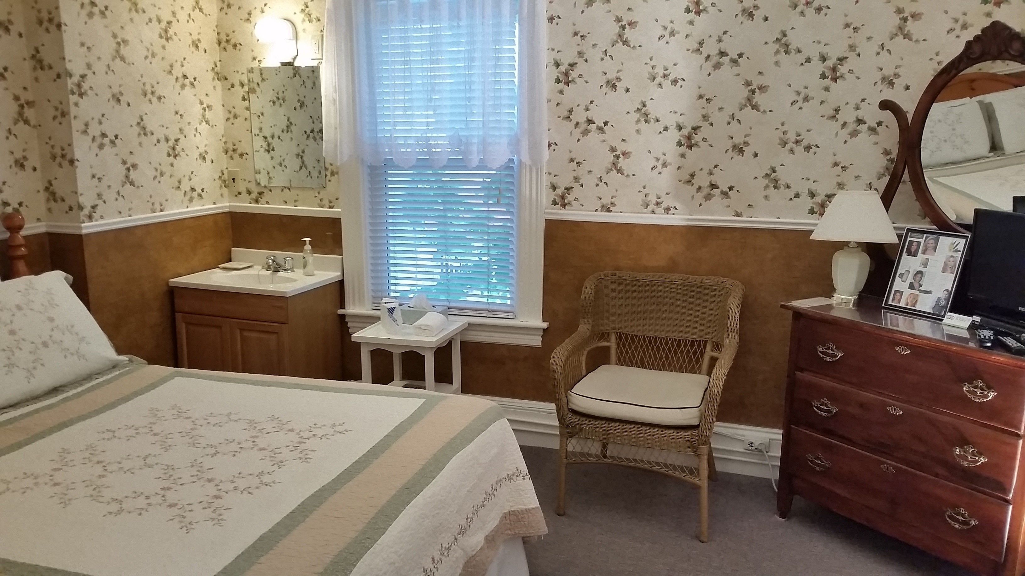 Un dormitorio con cama, una silla, cómoda, espejo, ventana con persianas y una mesita de noche con lámpara.