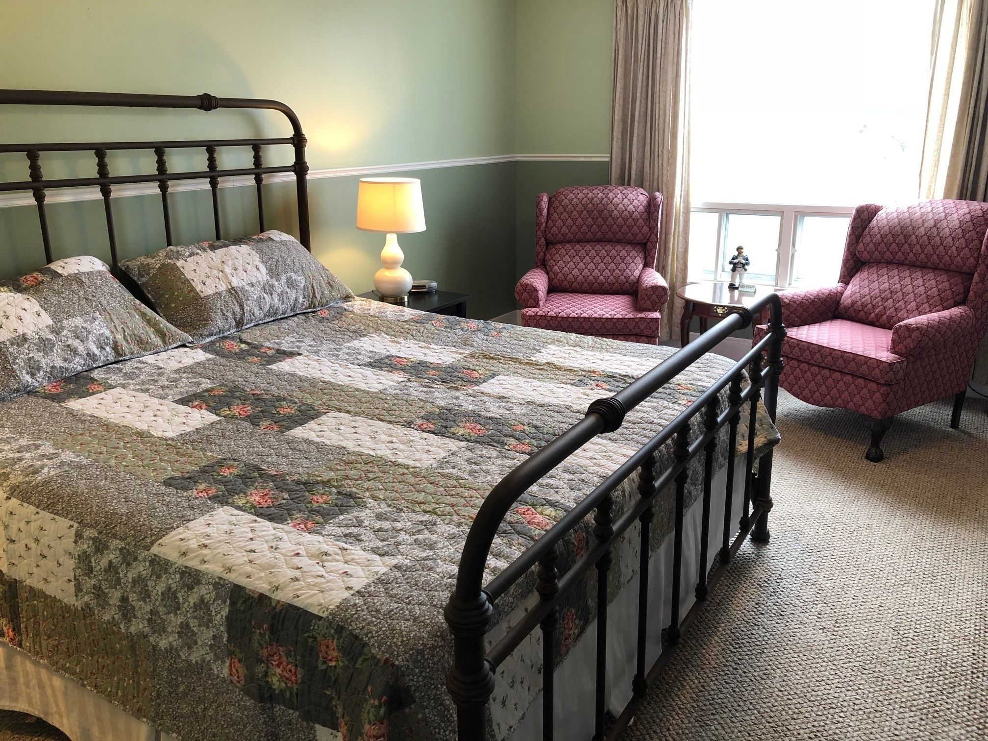 Una cama bien hecha con colcha floral, una lámpara de noche, dos sillones estampados y un ventanal con cortinas.
