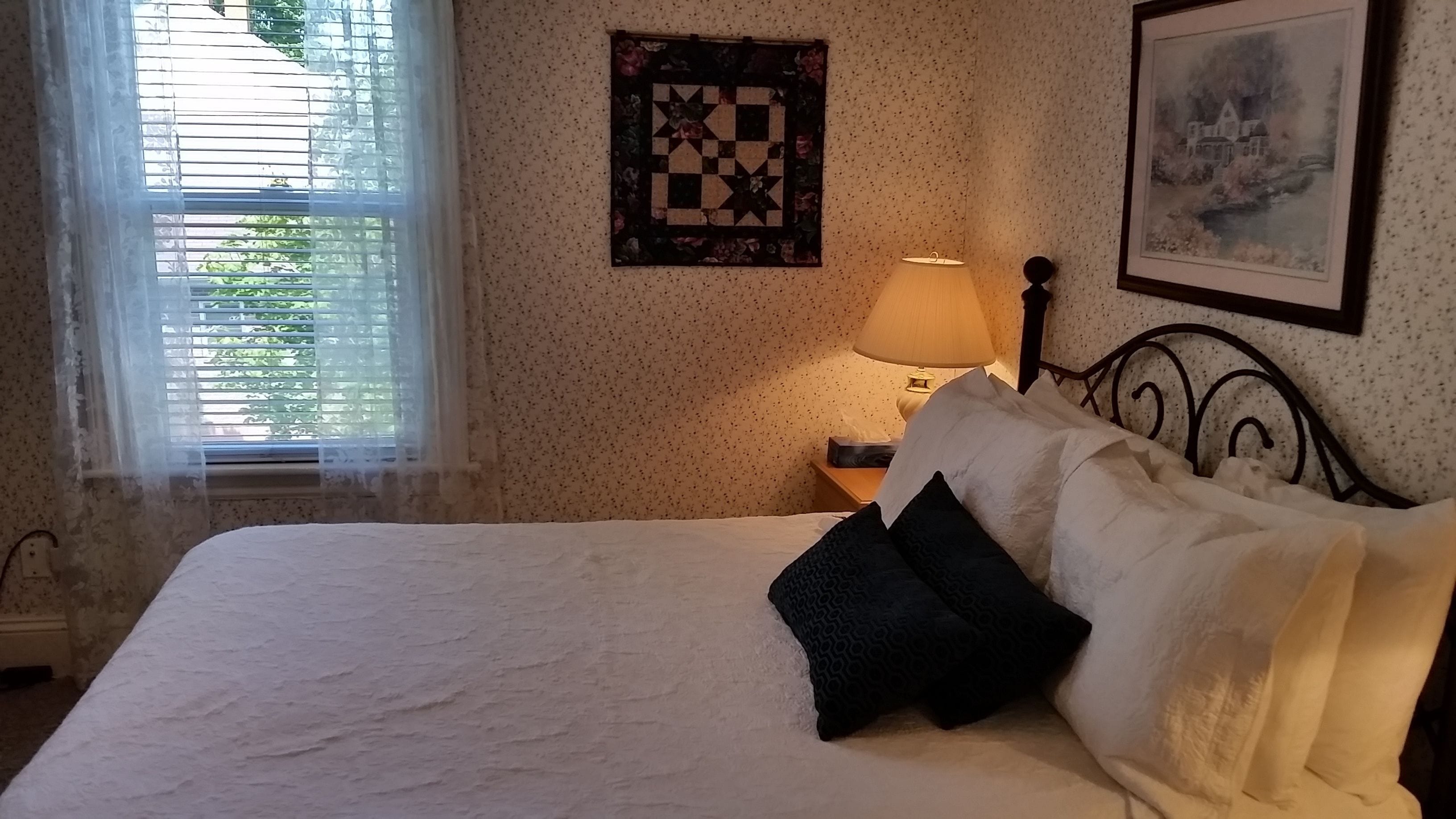 Een net opgemaakt bed met witte en zwarte kussens, een gewatteerde wanddecoratie, een ingelijst schilderij en een bedlampje.