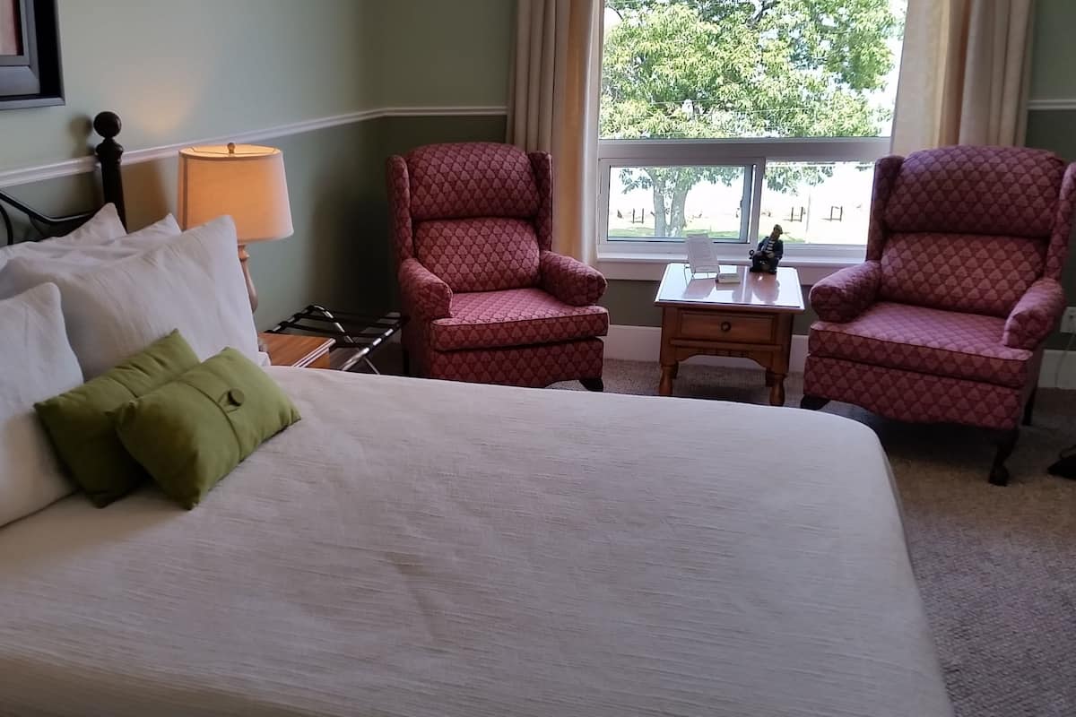 A hotel room with a large bed, two red patterned armchairs, a small table, and a window with a view of trees.