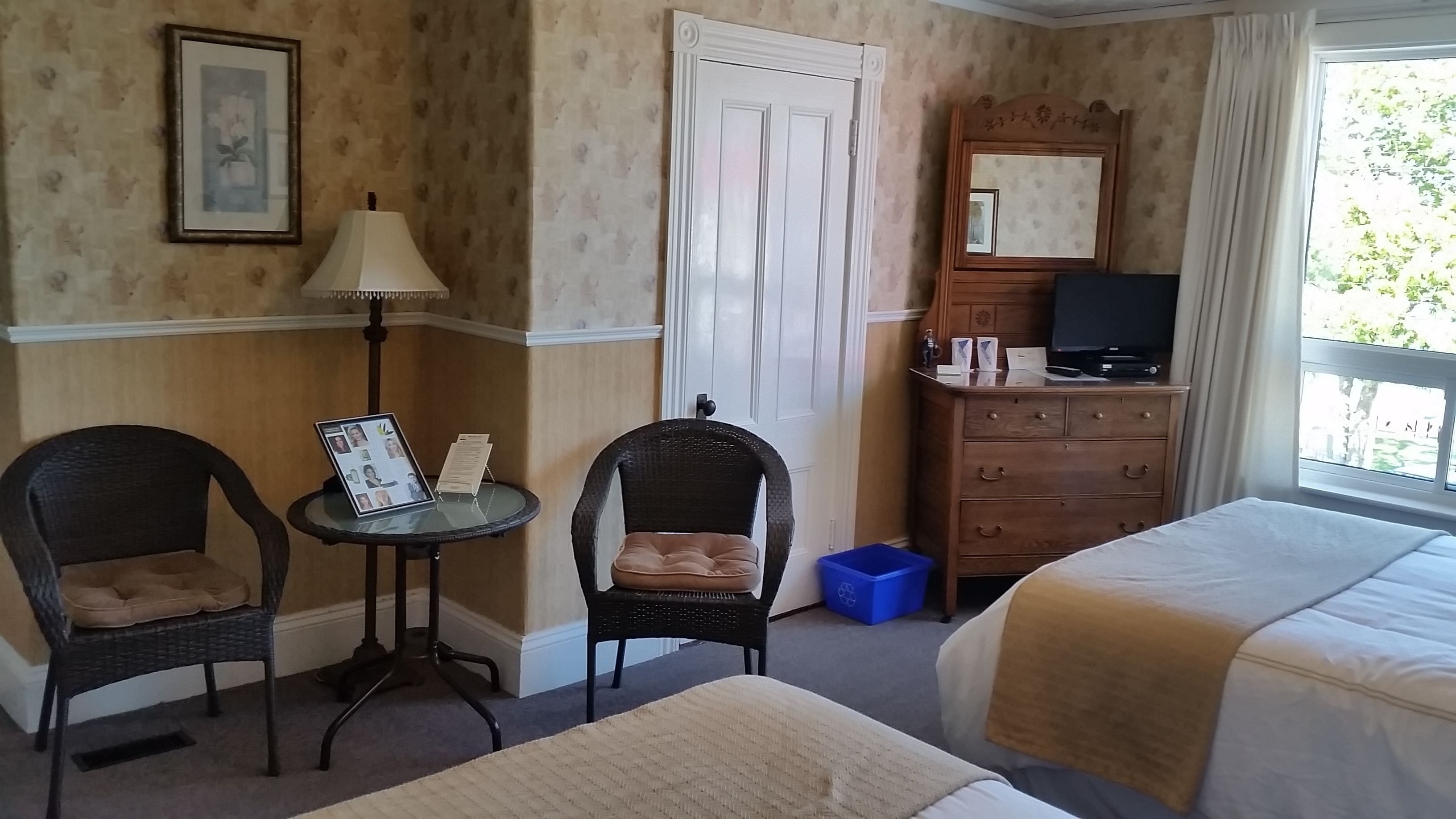 Hotelkamer met twee eenpersoonsbedden, een tafeltje met een lamp, een stoel, een dressoir met een televisie en een raam met uitzicht op groen.
